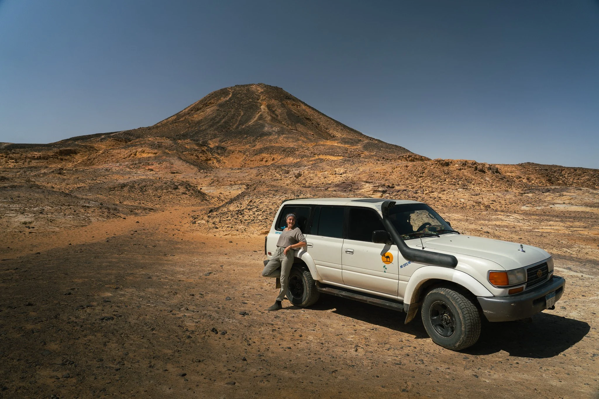 A person leaning against a white SUV in a desert landscape with a volcanic mountain in the background.