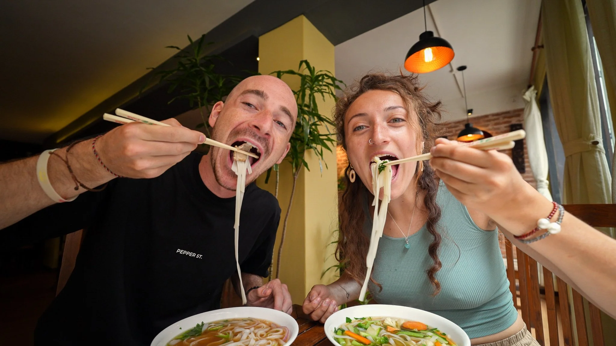 Two people enjoying a meal of noodles and soup at a restaurant, smiling and looking at the camera.