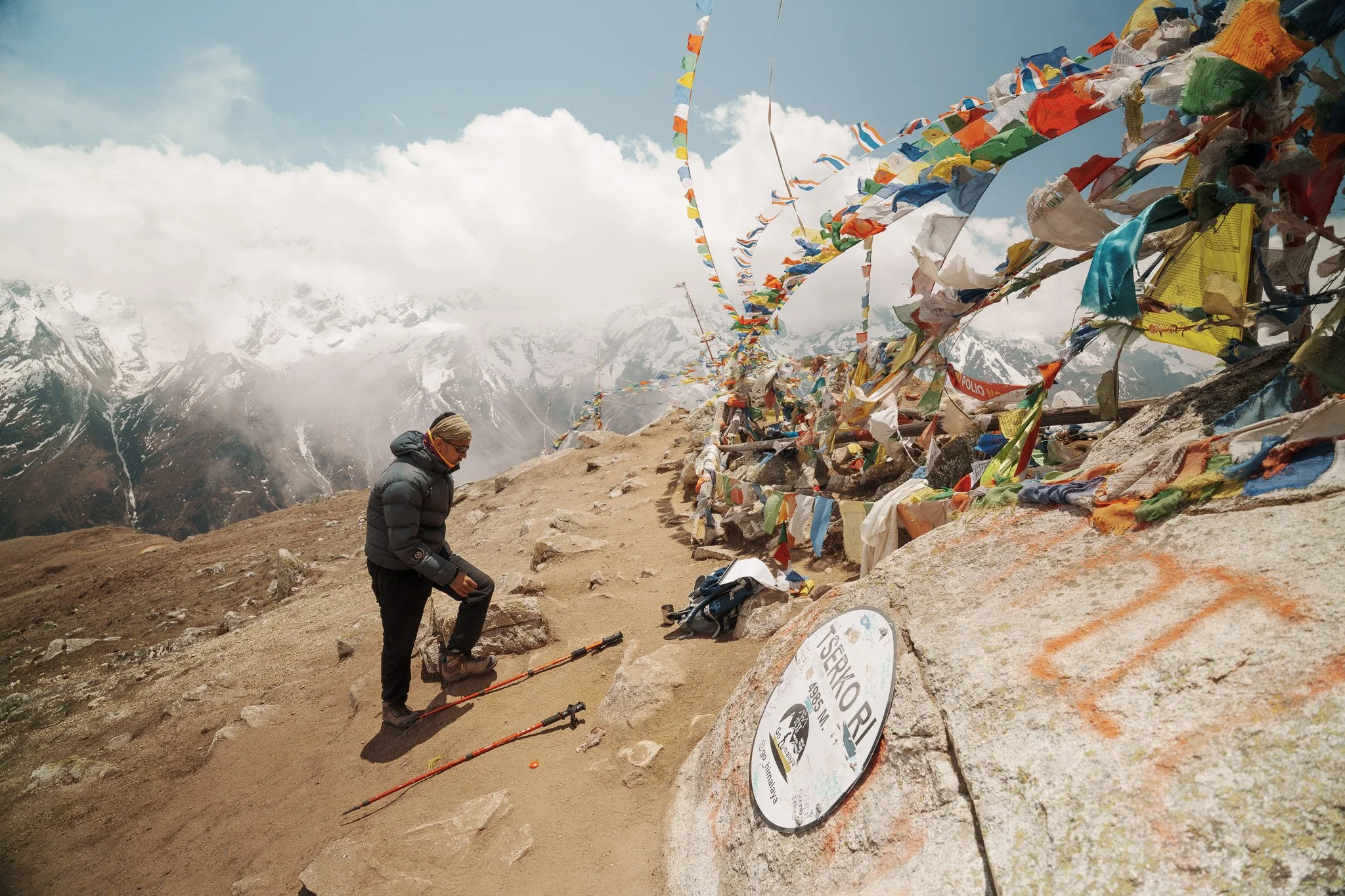 A hiker ascending a mountain trail with prayer flags lining the right side, snow-capped peaks in the background, and a mountain sign in the foreground reading 'Tserkio RI, 4055 m'.