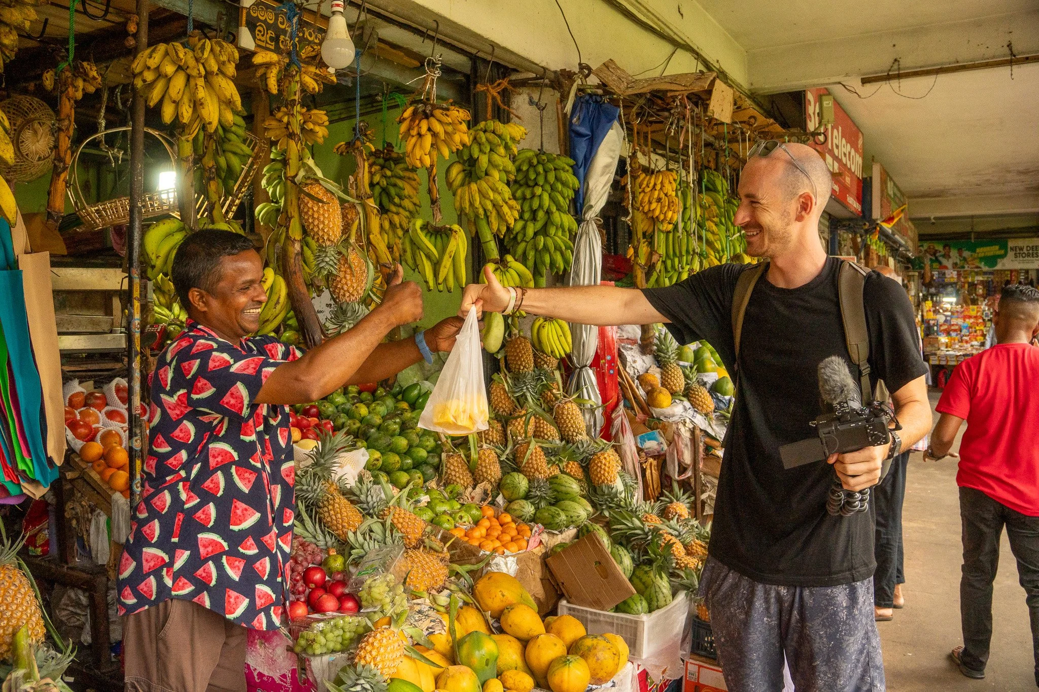 A man buying fruit from a local vendor at a market. The vendor is smiling and handing the man a plastic bag of fruit. The market stall is filled with various fruits, including bananas, pineapples, and other tropical fruits.