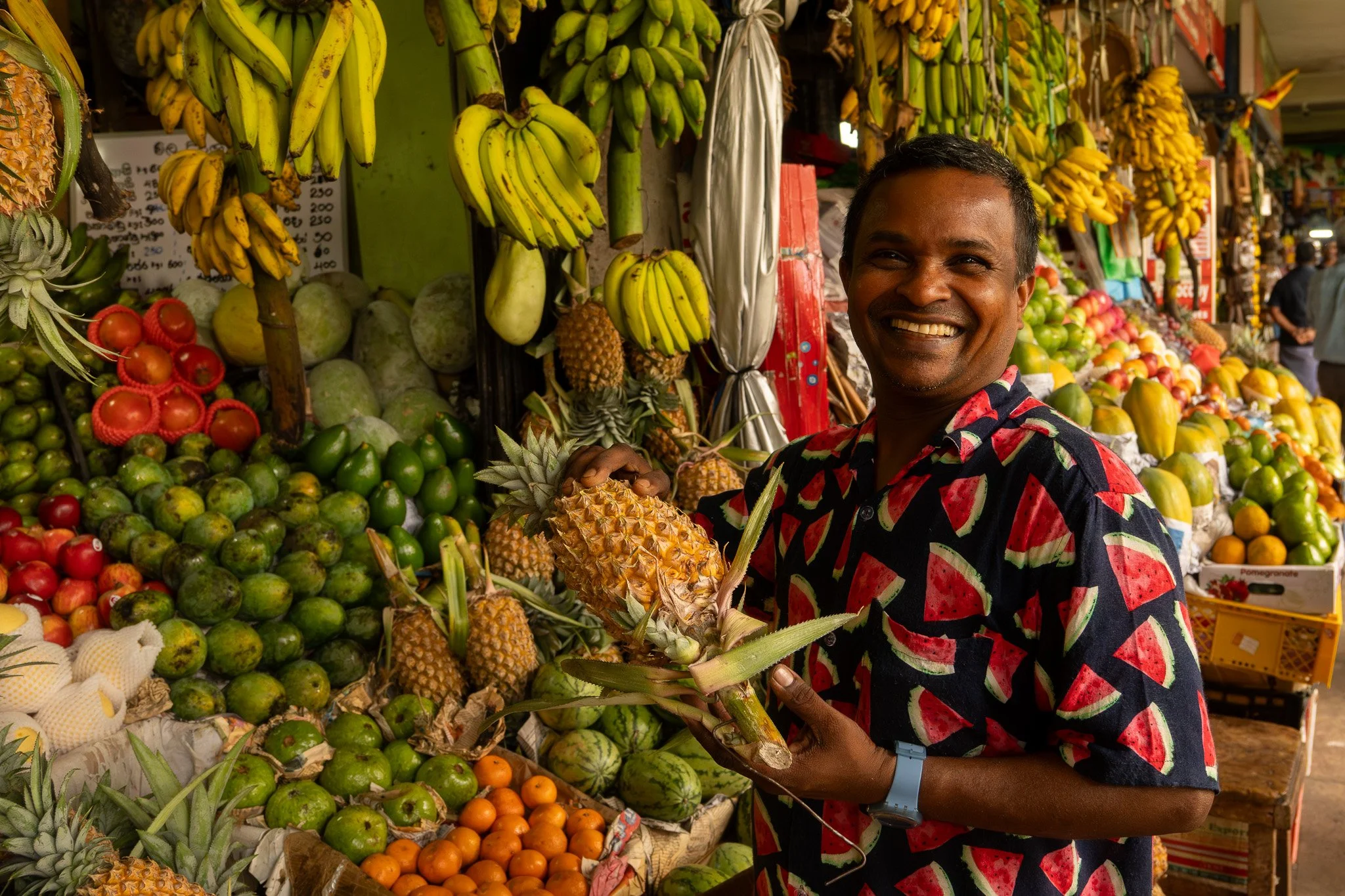 A man smiling while holding a pineapple at a colorful fruit market with bananas, watermelons, papayas, and other tropical fruits.