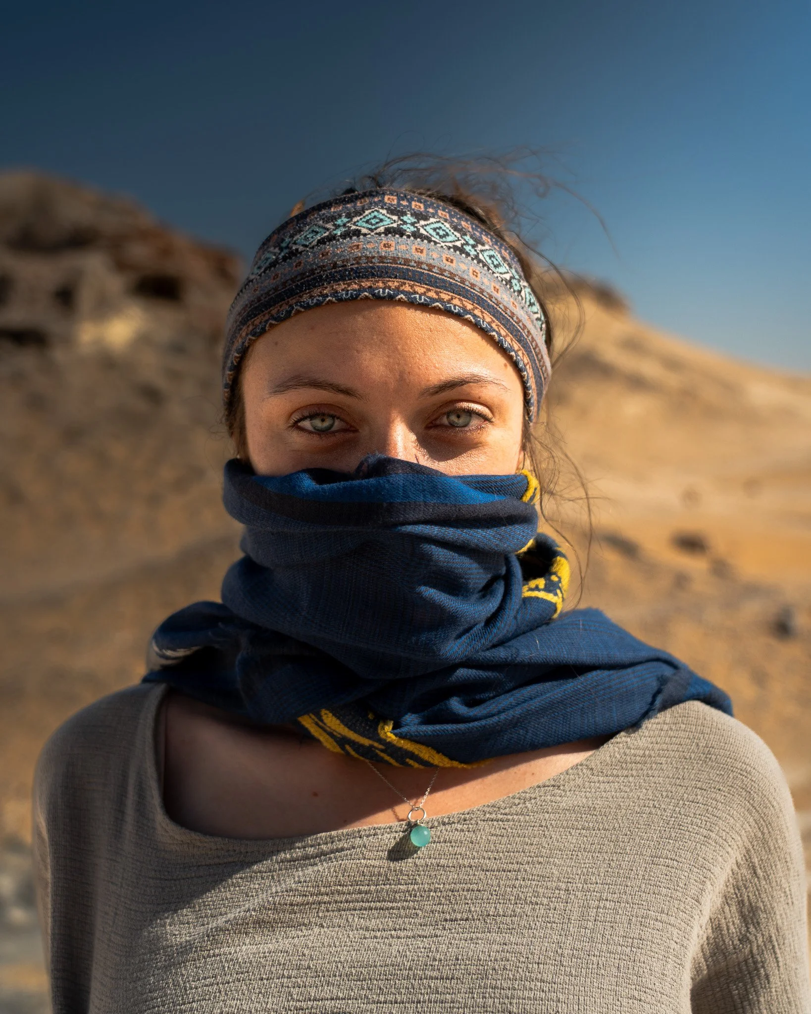 A woman with blue eyes and light skin, wearing a patterned headband and a blue neck scarf, standing outdoors in a desert landscape under a clear sky.