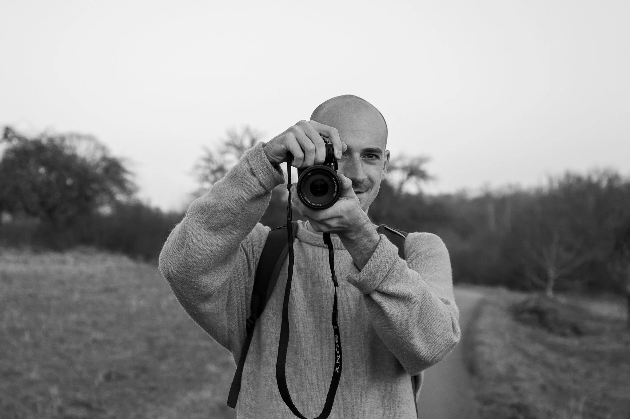 A man with a shaved head takes a photograph outdoors with a camera, standing on a grassy path with trees in the background, in black and white.