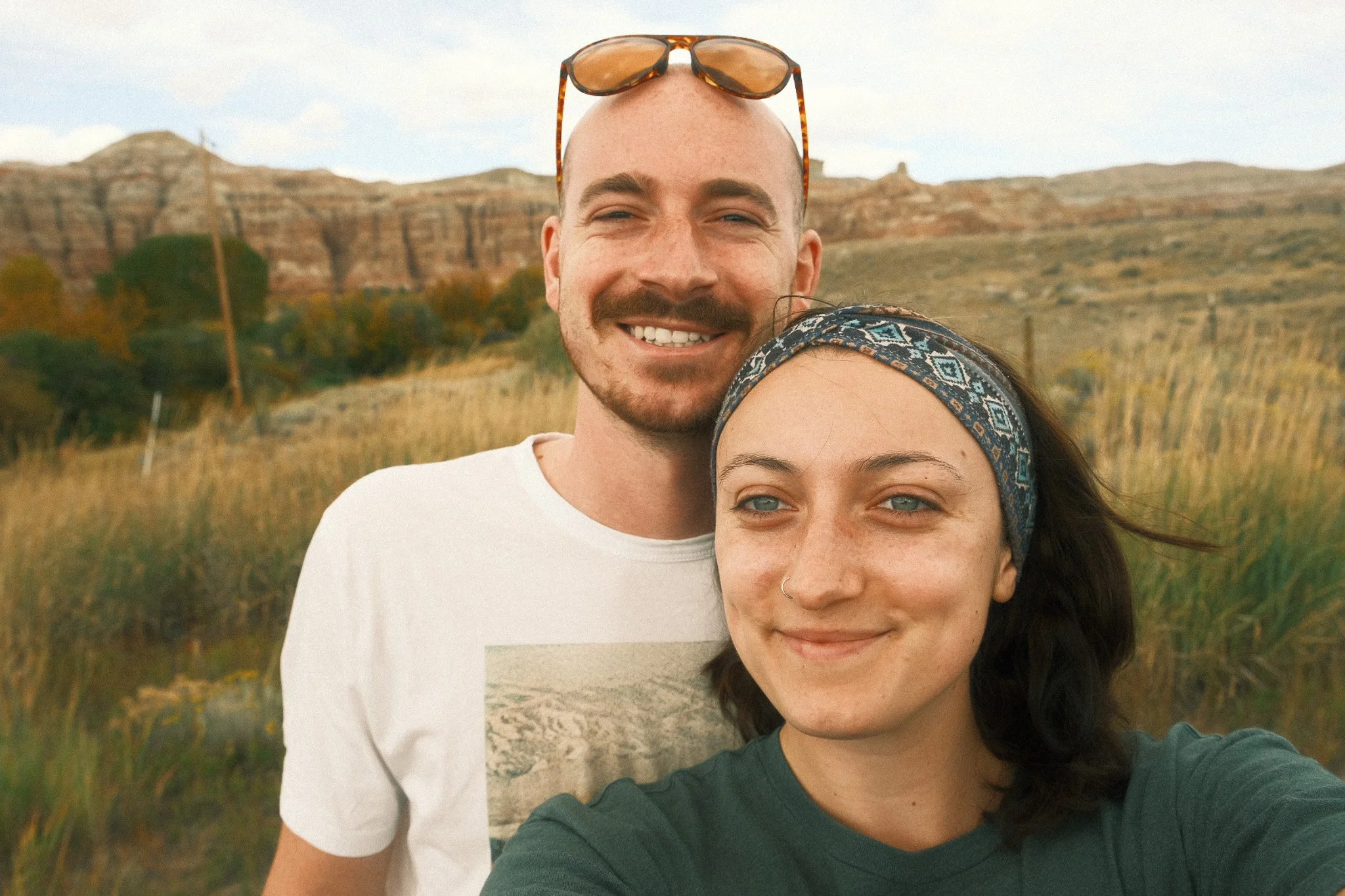 A young man with a beard and sunglasses on his head standing next to a young woman with a headband, both smiling outdoors with a natural landscape of grass, trees, and rocky hills in the background.