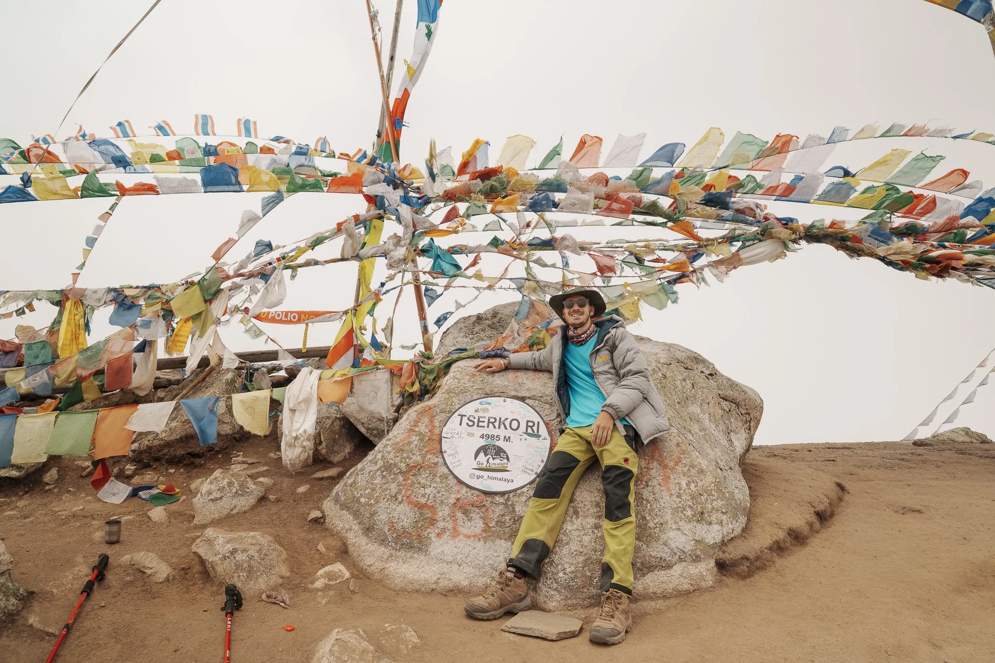 A man is sitting on a large rock at the summit of Tserko Ri mountain, surrounded by colorful prayer flags. He is wearing outdoor hiking gear, sunglasses, and a hat, smiling at the camera. The summit marker indicates an elevation of 4,985 meters.