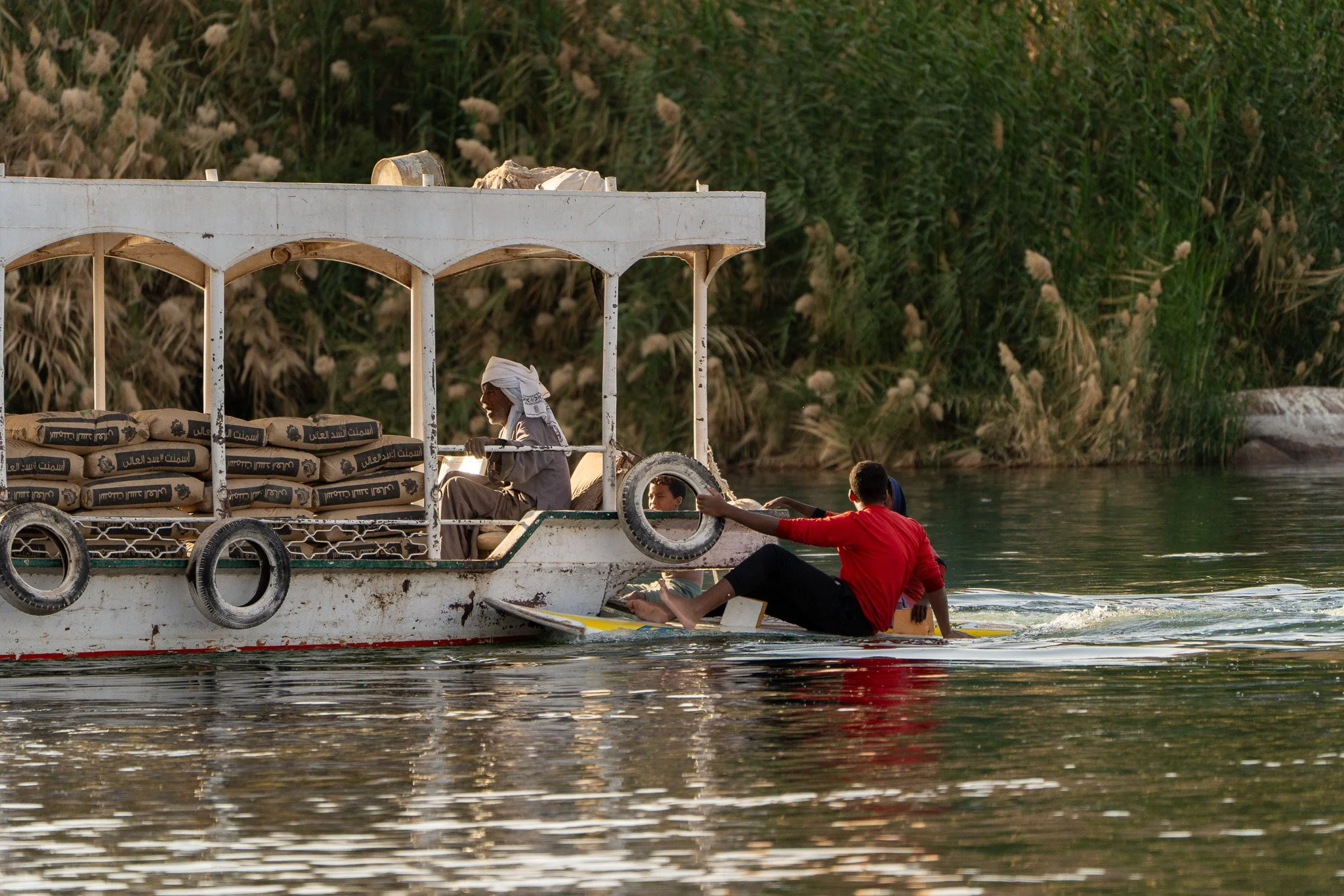 A boat on a river with a man in a red shirt holding onto the boat and sitting on a paddleboard, and a person inside the boat reading, with a background of green trees and tall grass.