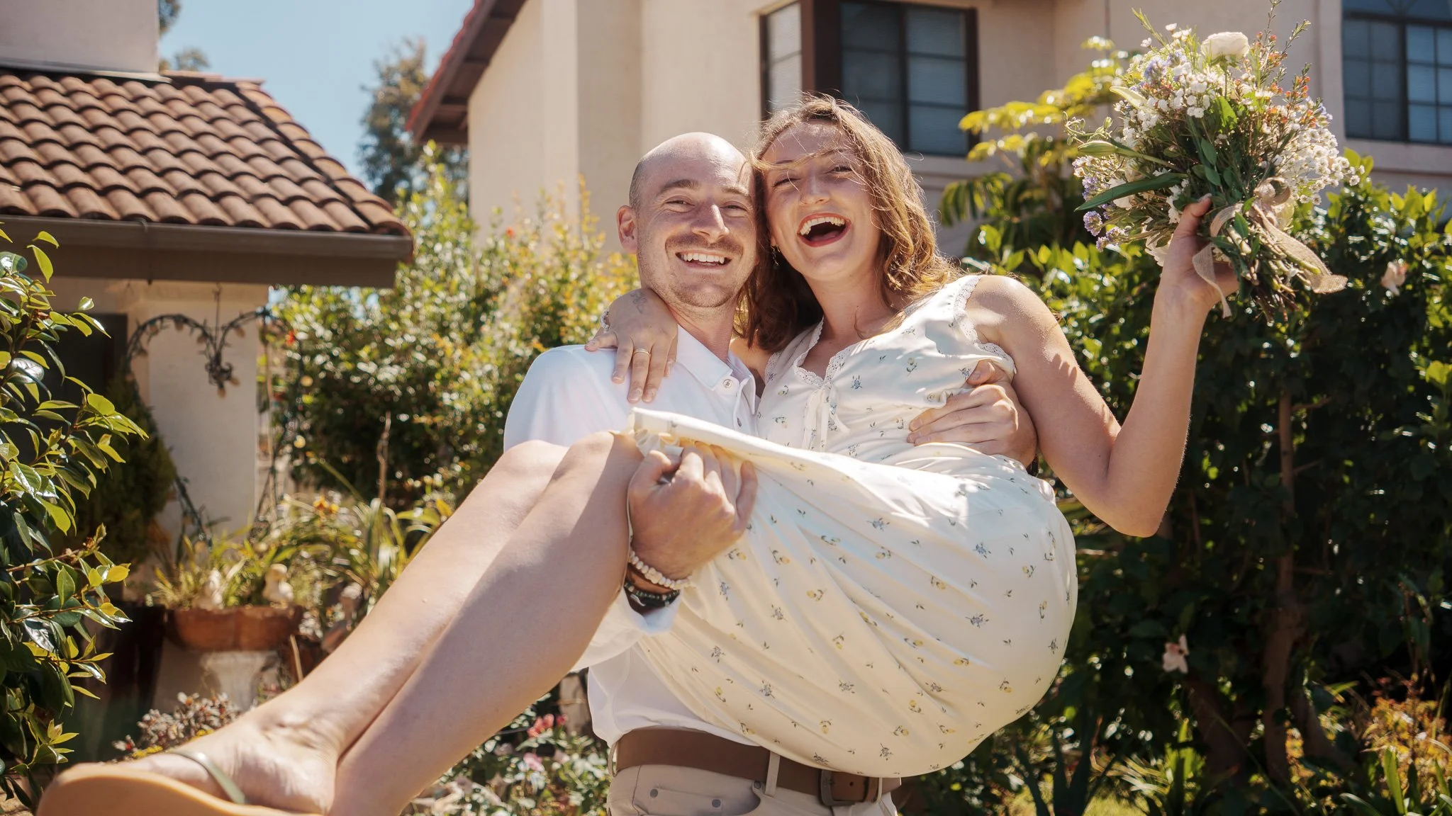 A couple smiling happily, with the man holding the woman in his arms, a woman holding a bouquet of flowers, standing outdoors in a garden with a house in the background on a sunny day.