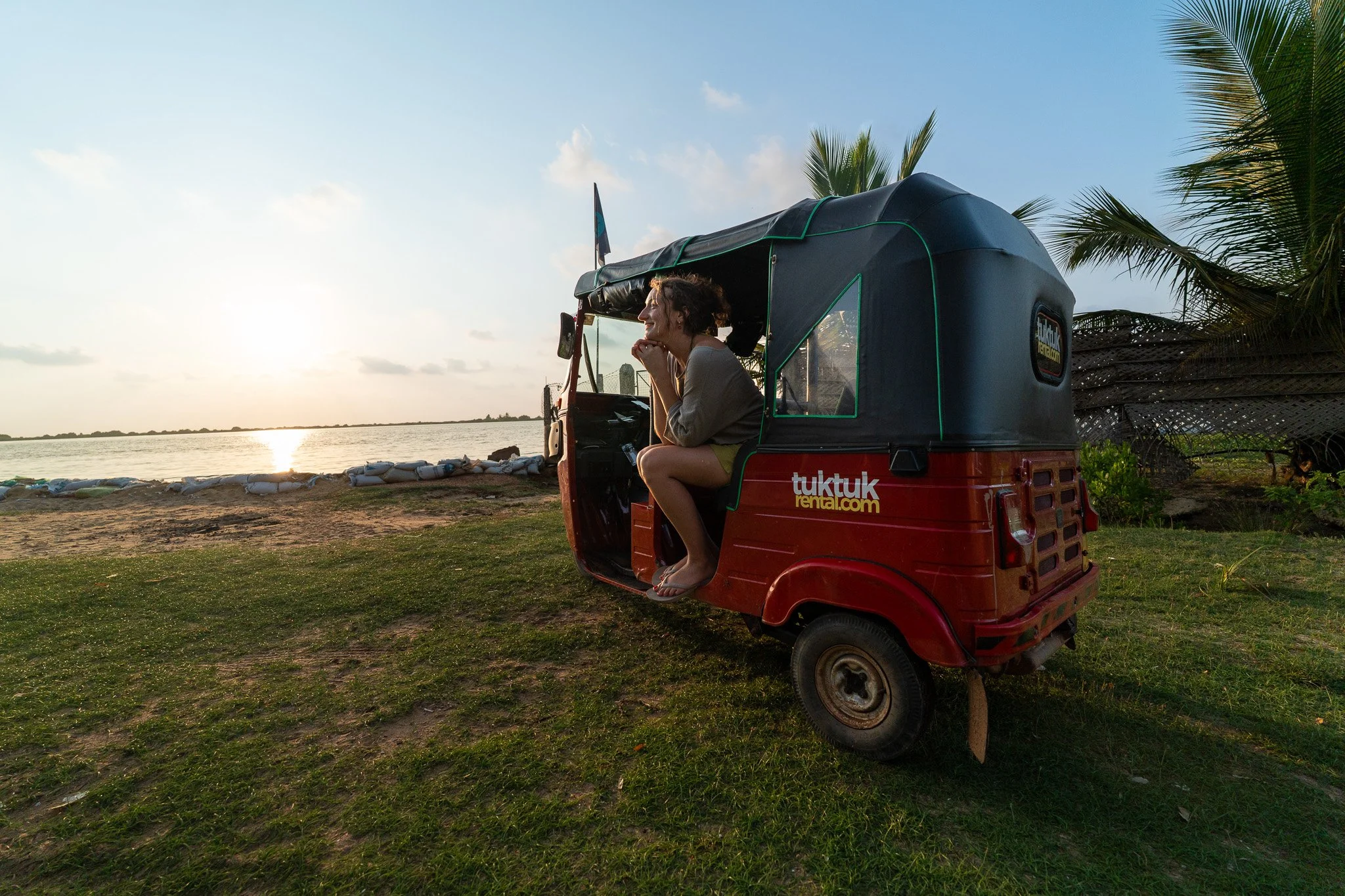 A woman sitting inside a red tuk tuk parked on the grassy shore near a body of water during sunset.