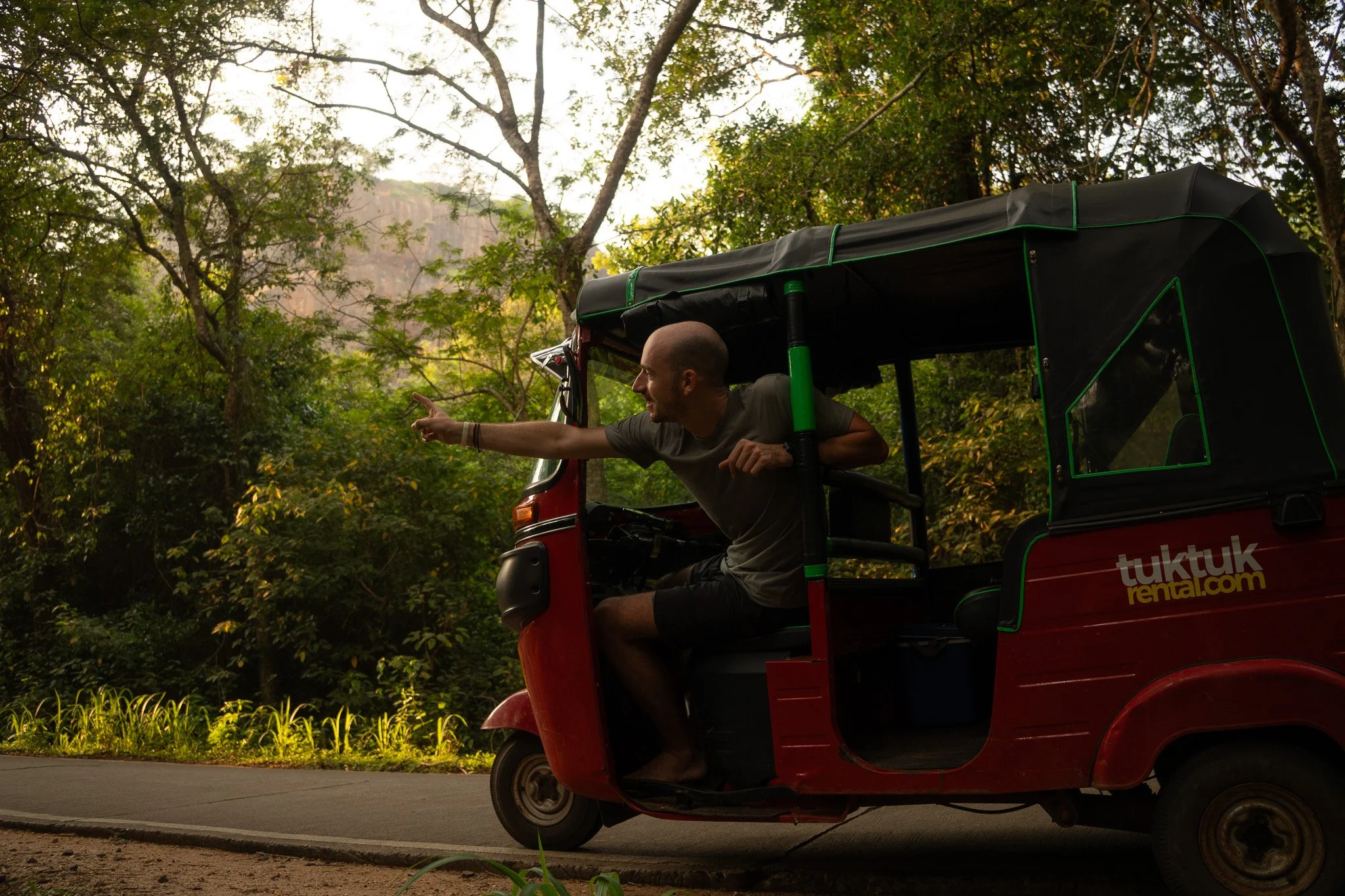 A man in a grey t-shirt and shorts inside a red tuk-tuk, reaching out and pointing at something on the side of the road in a lush forest setting.