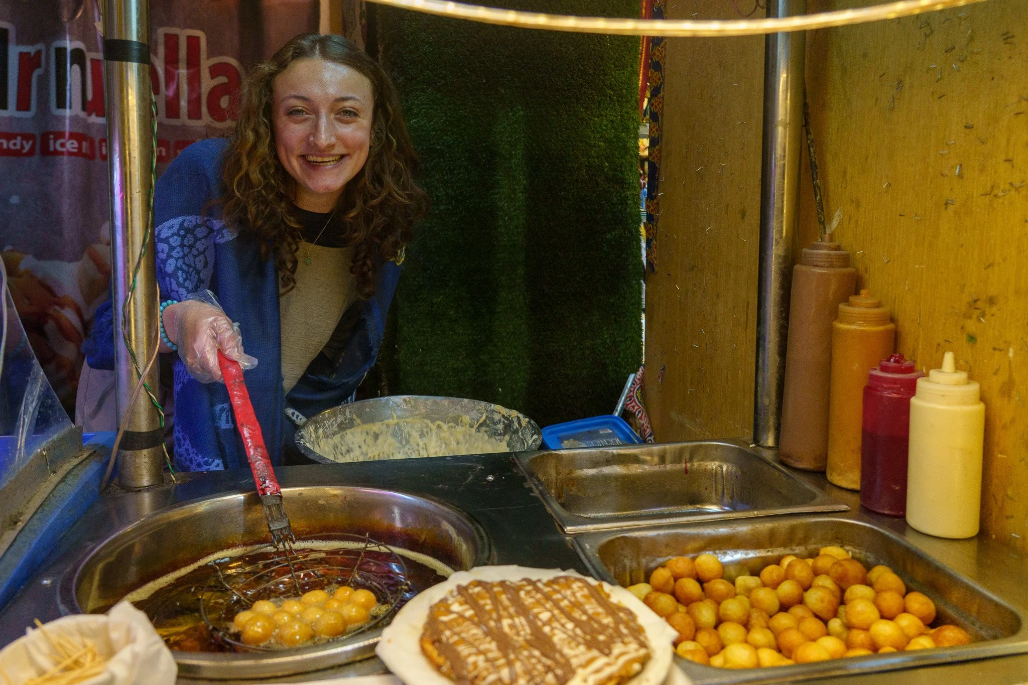 Young woman cooking at a food stall, frying small round fried foods in hot oil, with bottles of condiments and a plate of fried dough nearby.