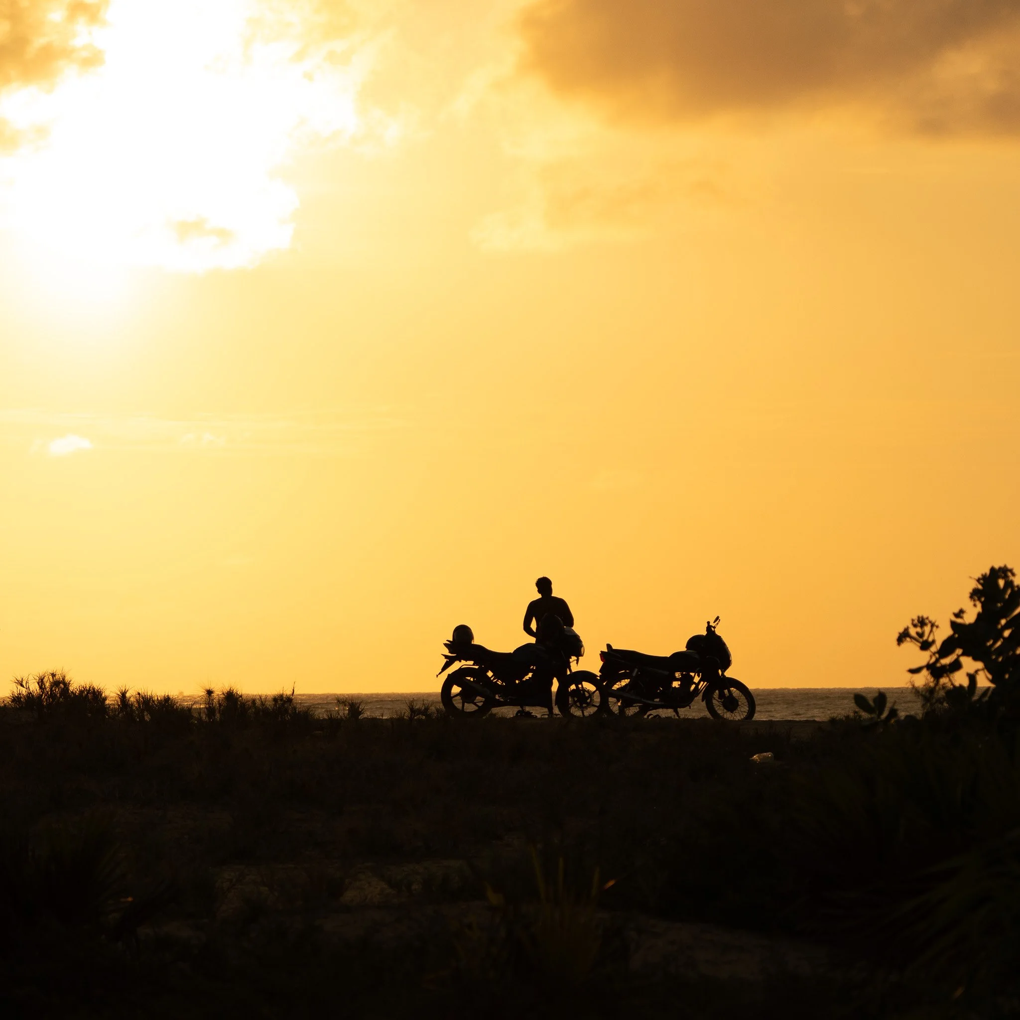 Silhouettes of two motorcycles and a person standing between them at sunset, with a yellow-orange sky in the background.