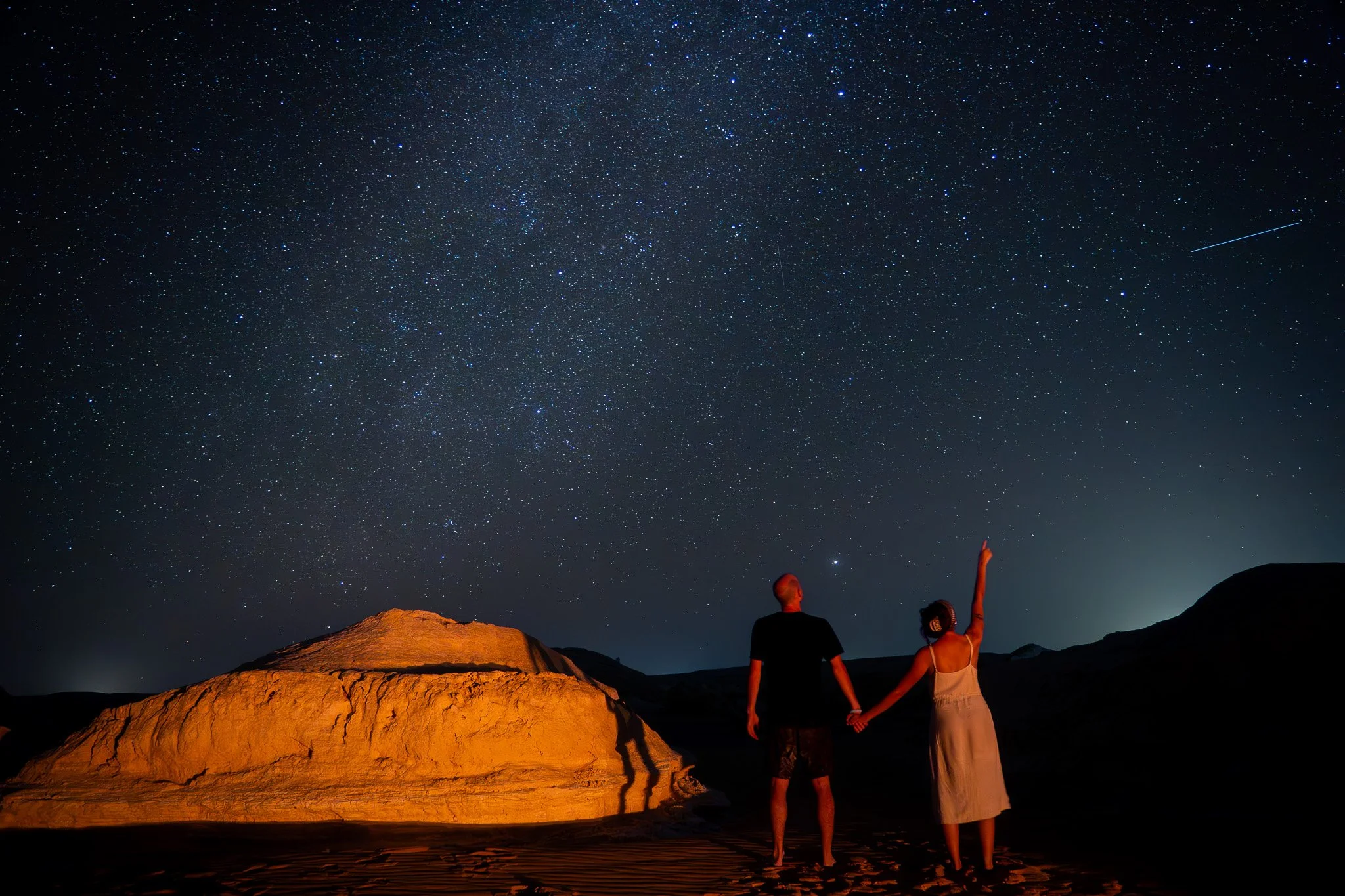 A couple holding hands and watching the night sky filled with stars and a shooting star in a desert landscape with illuminated rocks.