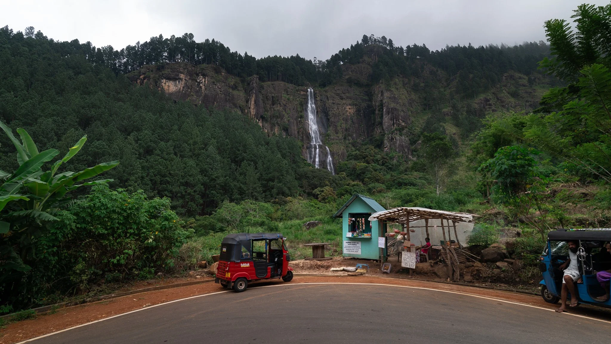 Scenic view of a waterfall flowing down a lush green mountainside, with small roadside vendors and tuk-tuk vehicles near the road in the foreground.