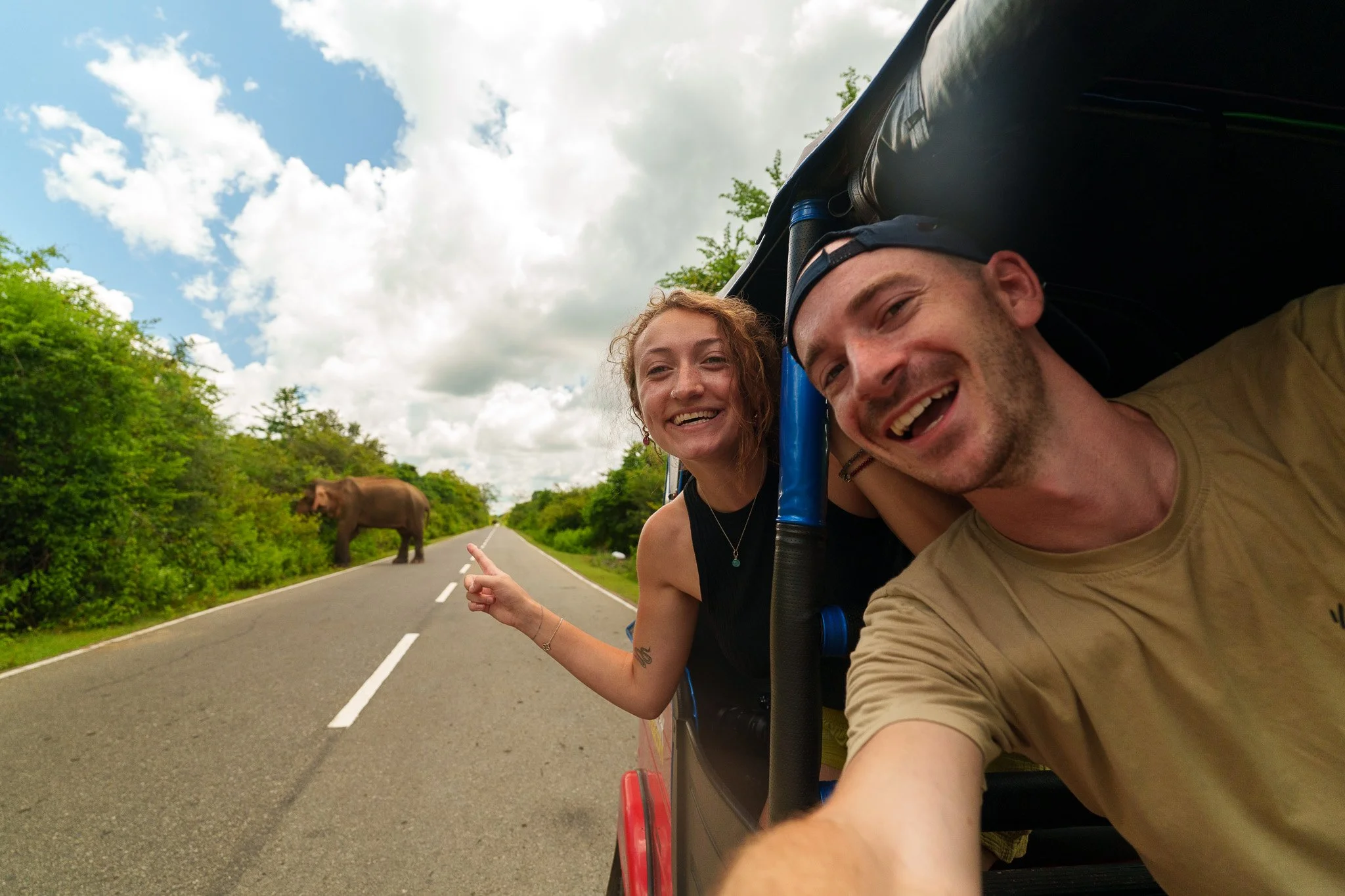 A young man and woman smiling and taking a selfie from a moving vehicle with an elephant on the road ahead, surrounded by greenery and cloudy sky.