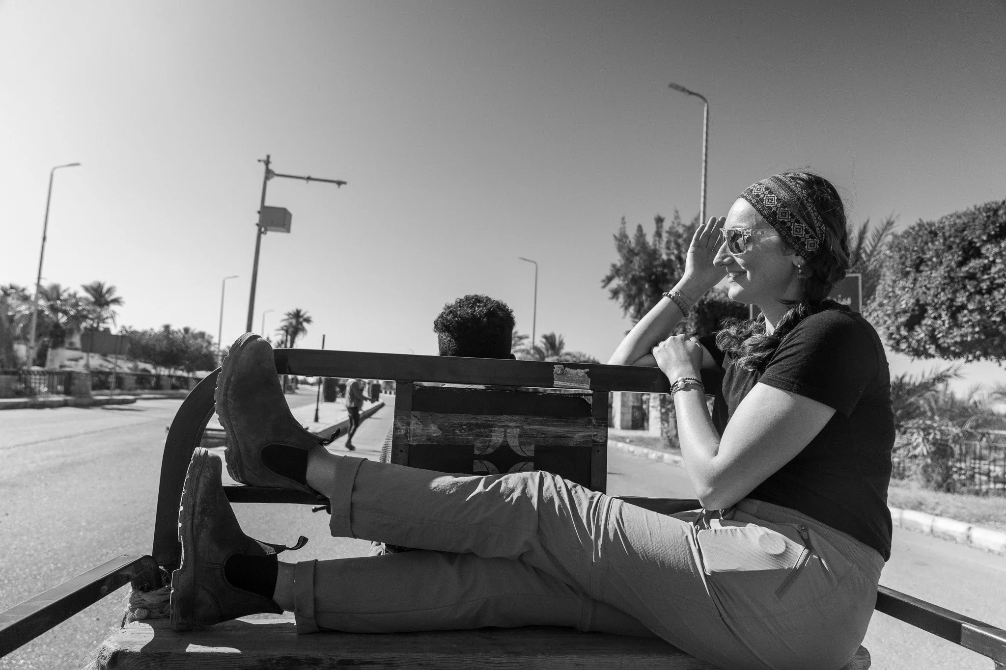 A woman sitting on a cart in a city street, wearing sunglasses, a bandana, and casual clothes, with her legs stretched out and smiling.