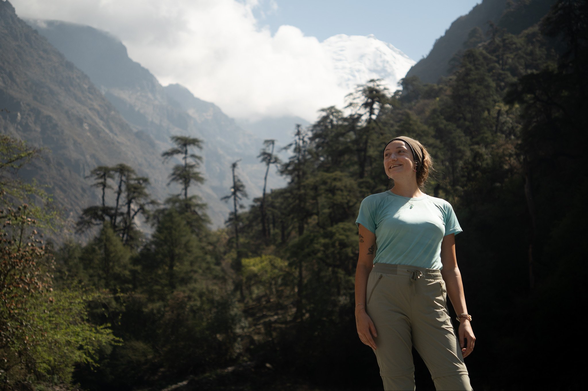 A woman smiles while standing in a mountainous forested area, with tall trees and snow-capped peaks in the background.