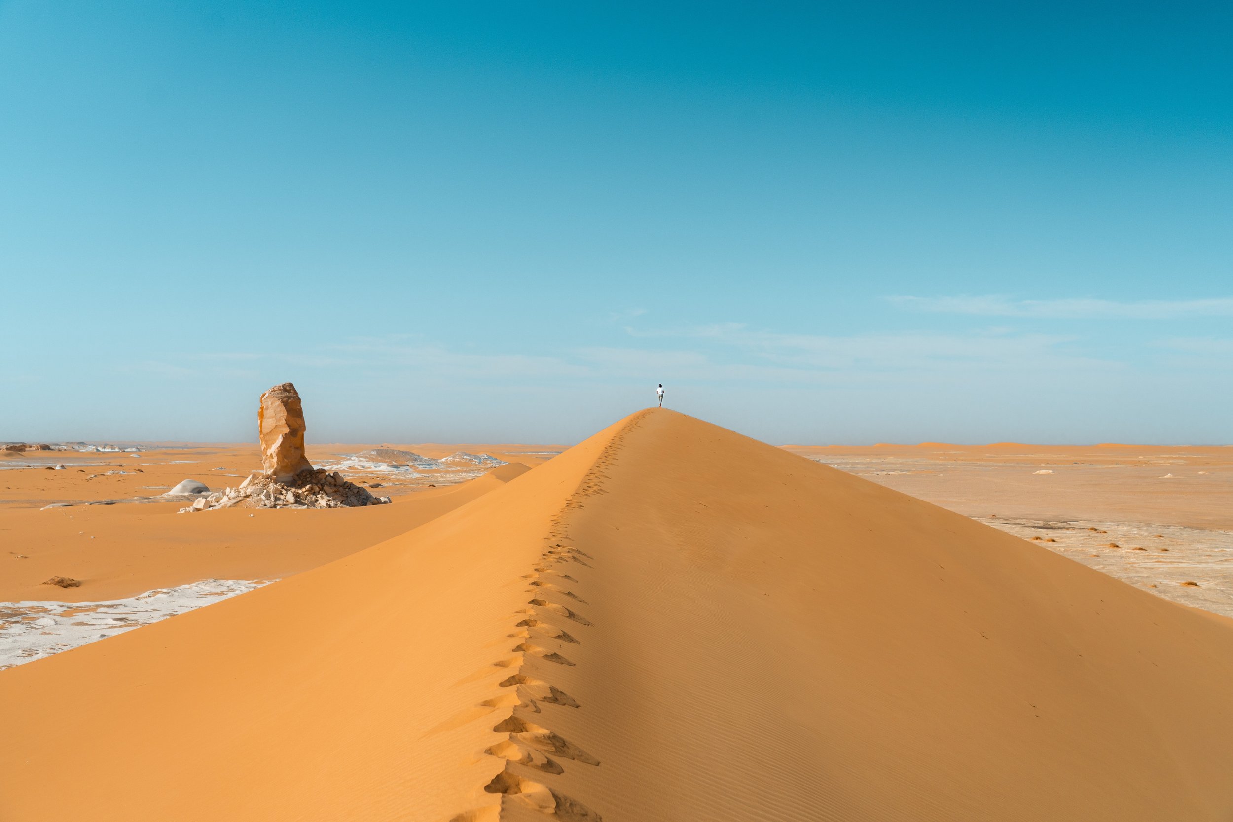 A photo of a person standing at the top of a sand dune in a desert, with a clear blue sky above and a rock formation in the background.