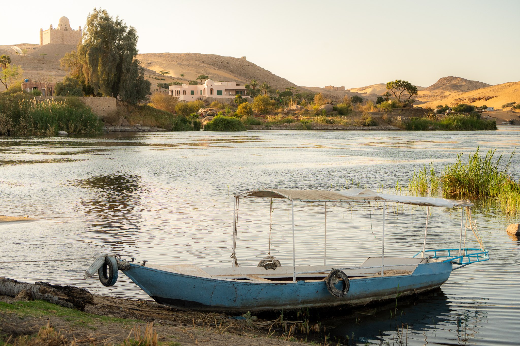 A blue boat resting on the riverbank with water, greenery, and houses on the opposite shore, with hills and a castle in the background at sunset.