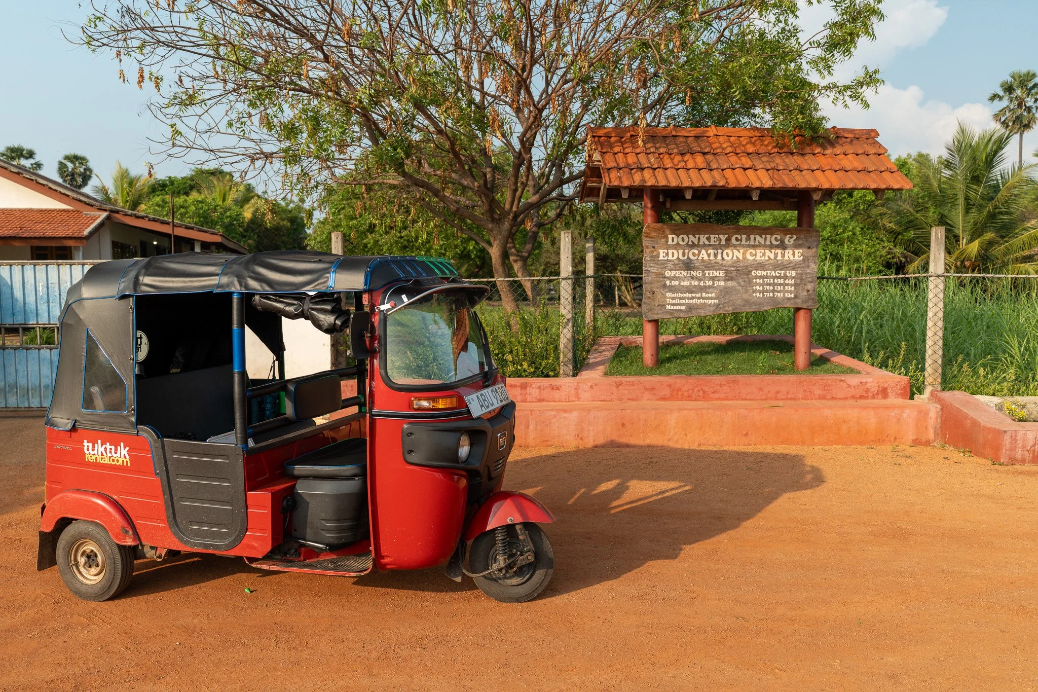 A red tuk-tuk parked on a dirt road in front of a sign for Donkey Clinic & Education Centre, with a tree, a fence, and some buildings in the background.