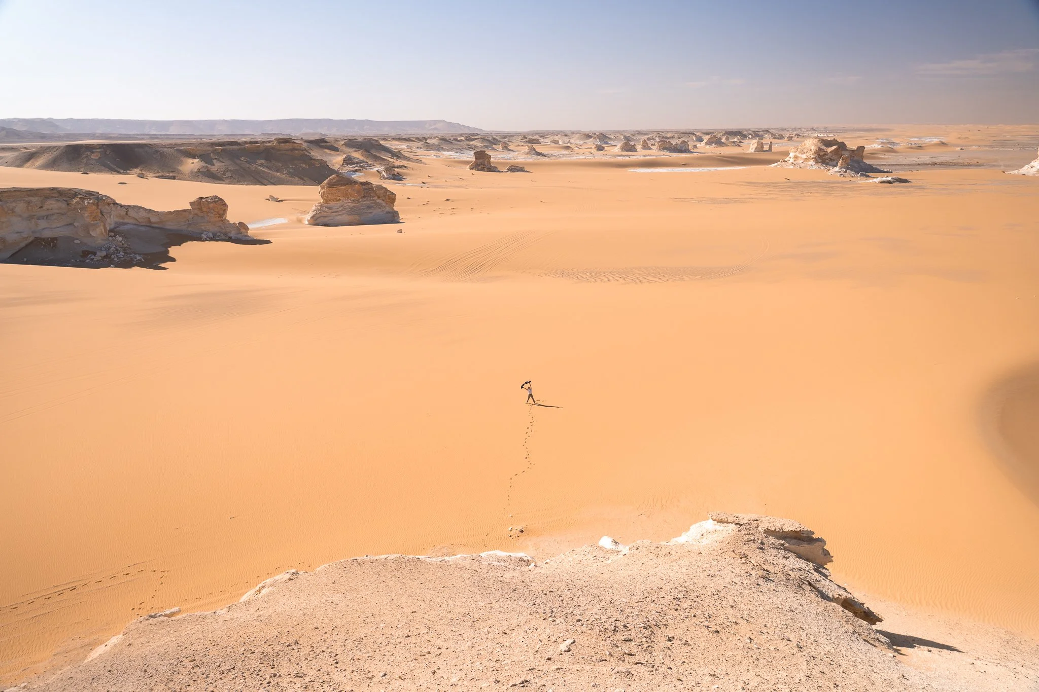 A person trail running in a vast desert with orange sand and scattered rock formations under a blue sky.