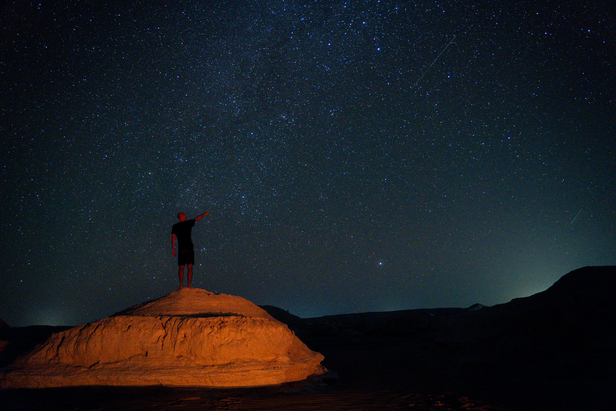 Person standing on a mound of earth pointing towards the starry night sky.