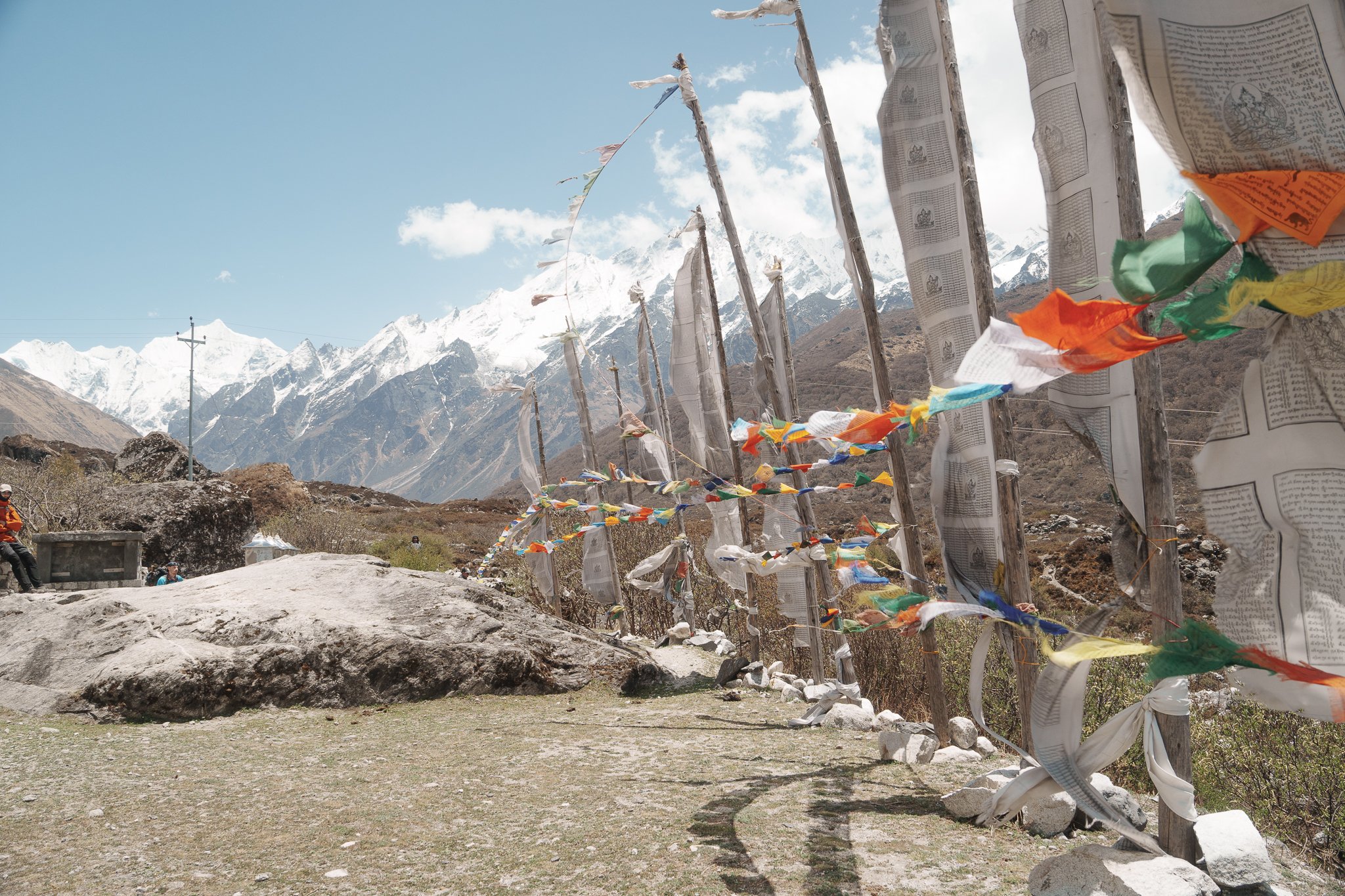 Colorful prayer flags attached to wooden poles on a cloudy mountain background with snow-capped peaks.