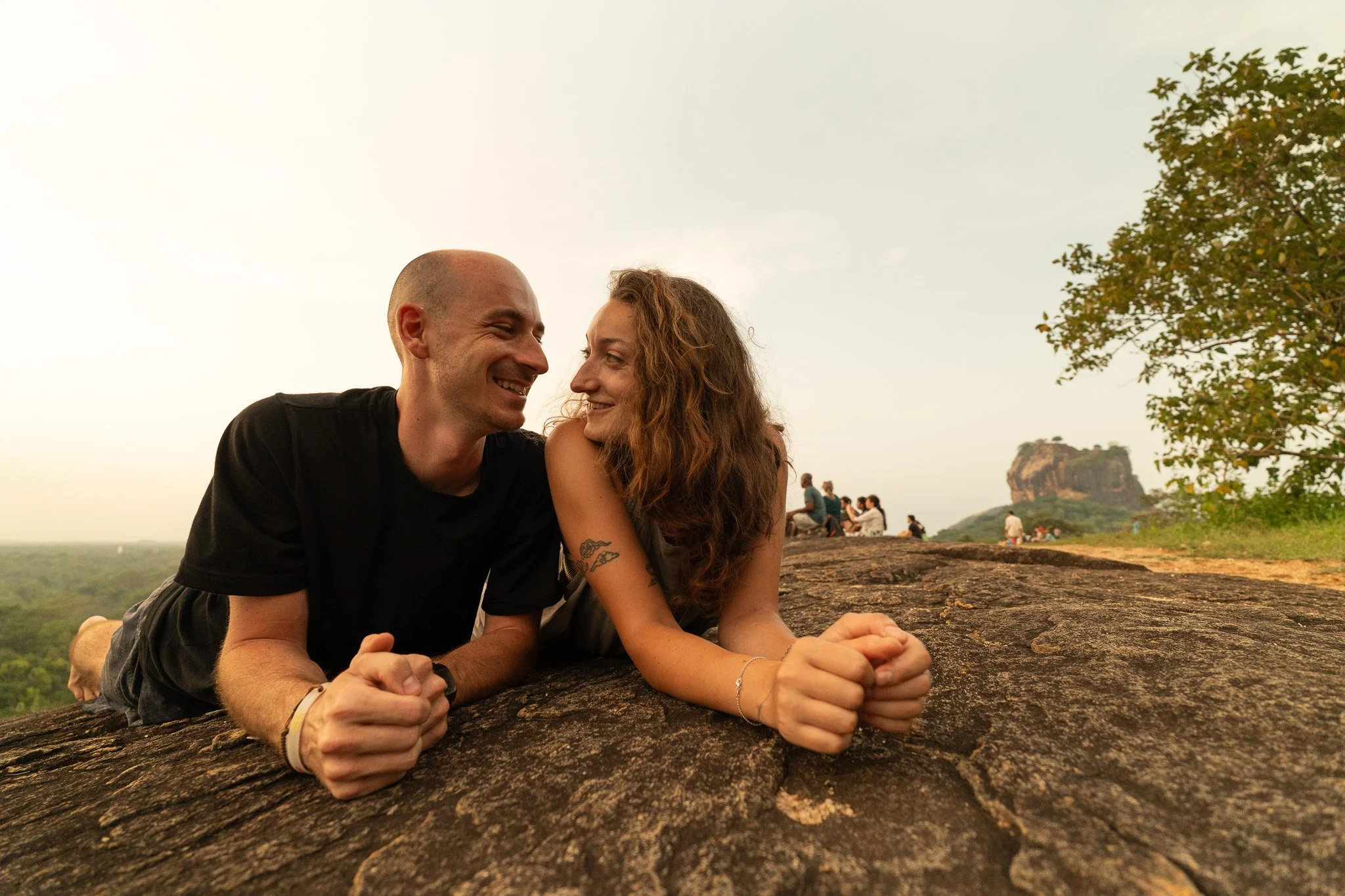 A couple lying on a large rock outdoors, smiling and looking at each other, with trees and a distant hill in the background.