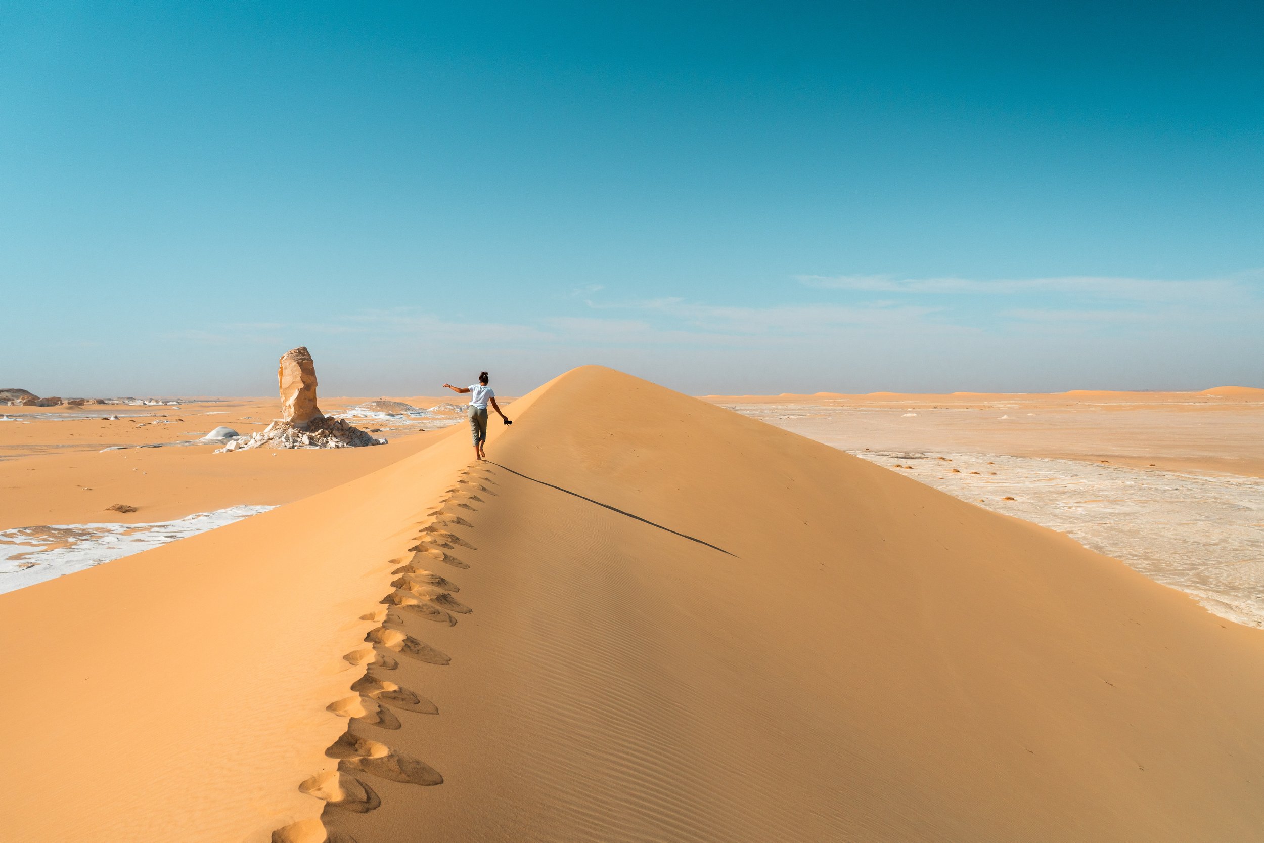 A person walking along the crest of a sand dune in a desert landscape, with footprints in the sand and a clear blue sky overhead.