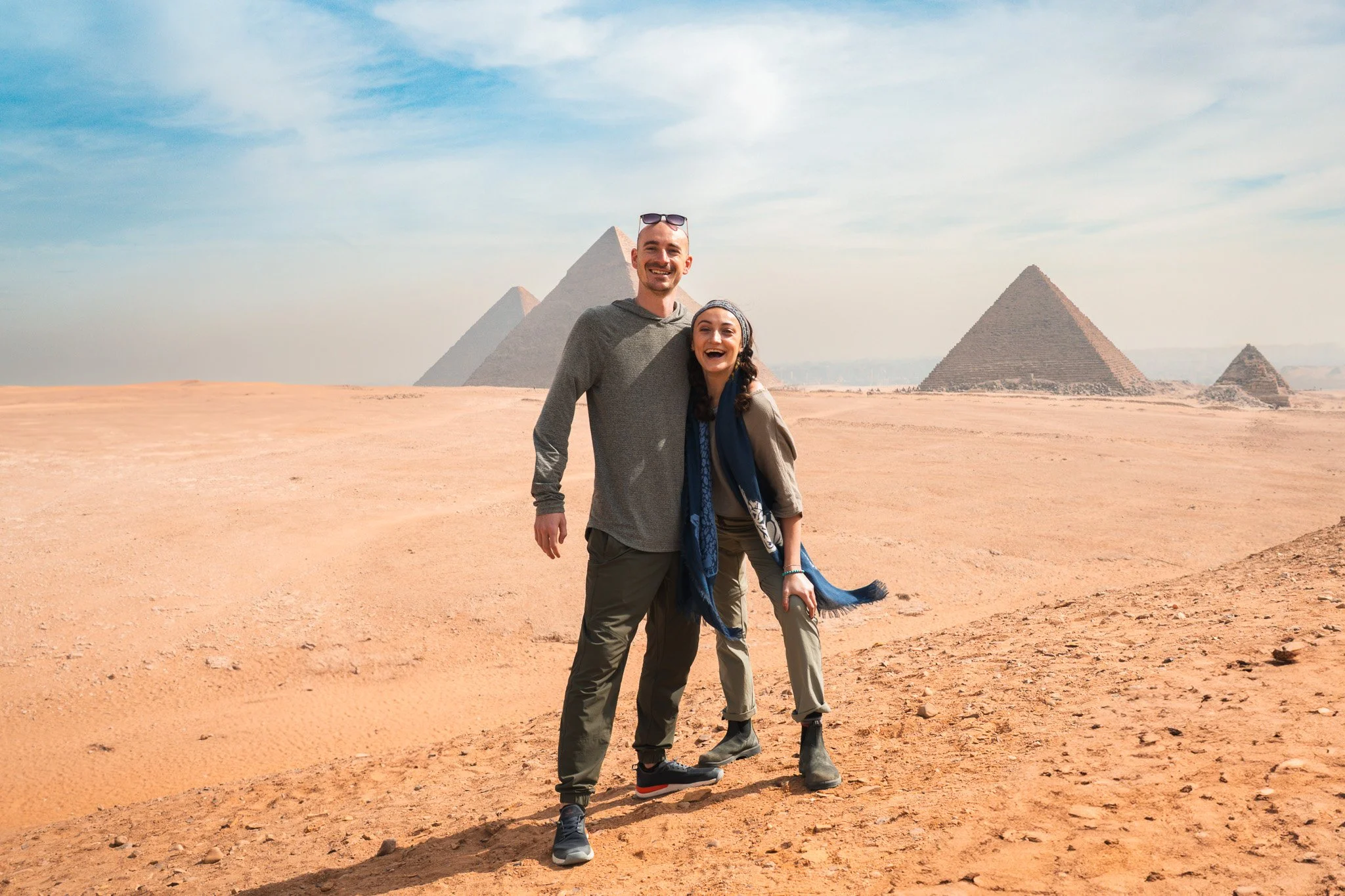 Two smiling tourists, a man and a woman, standing on sandy desert with pyramids in the background, likely in Egypt.