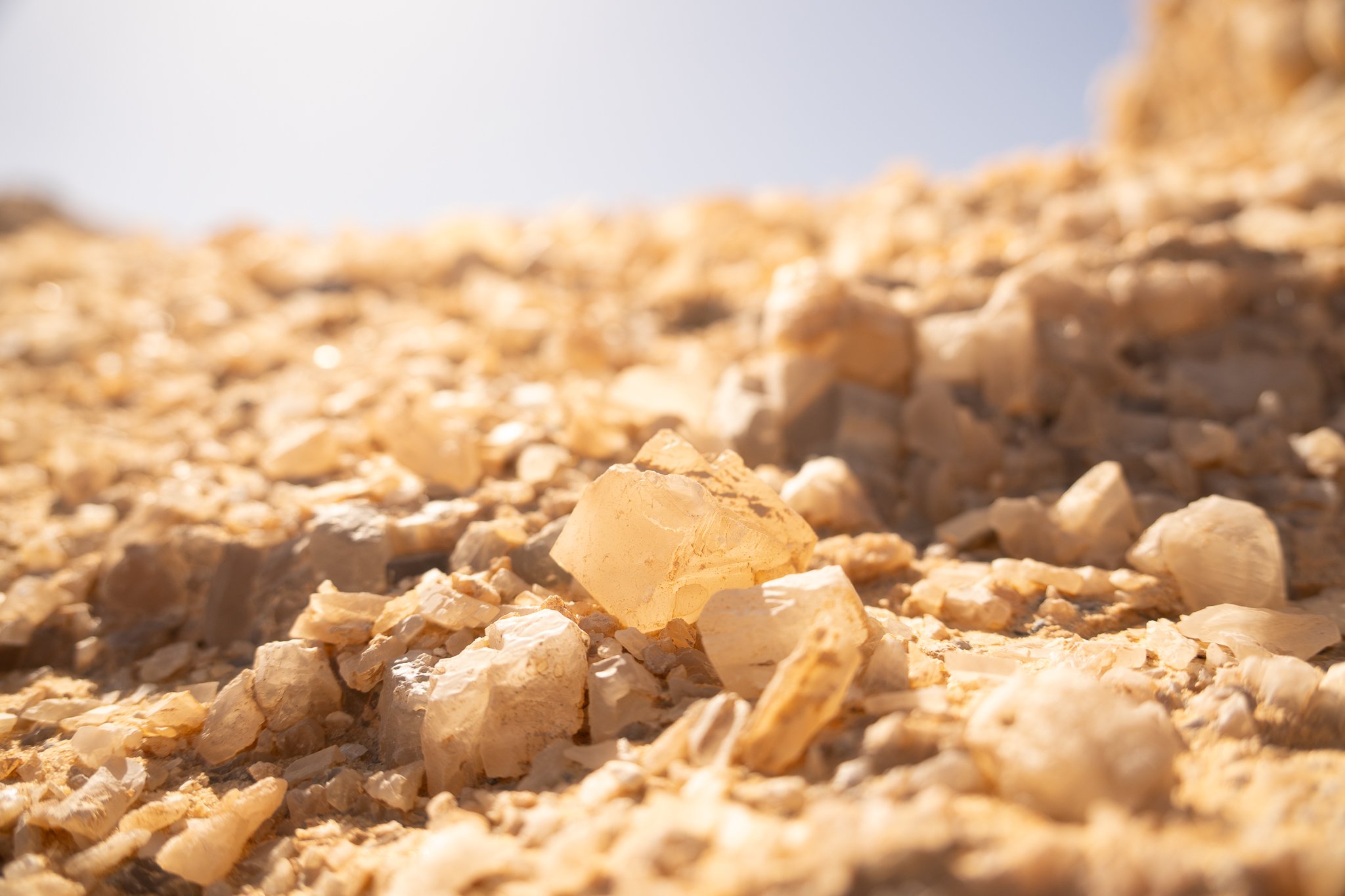 Close-up view of sand and small rocks, with lighter-colored stones in the foreground and a blurred background under sunny sky.