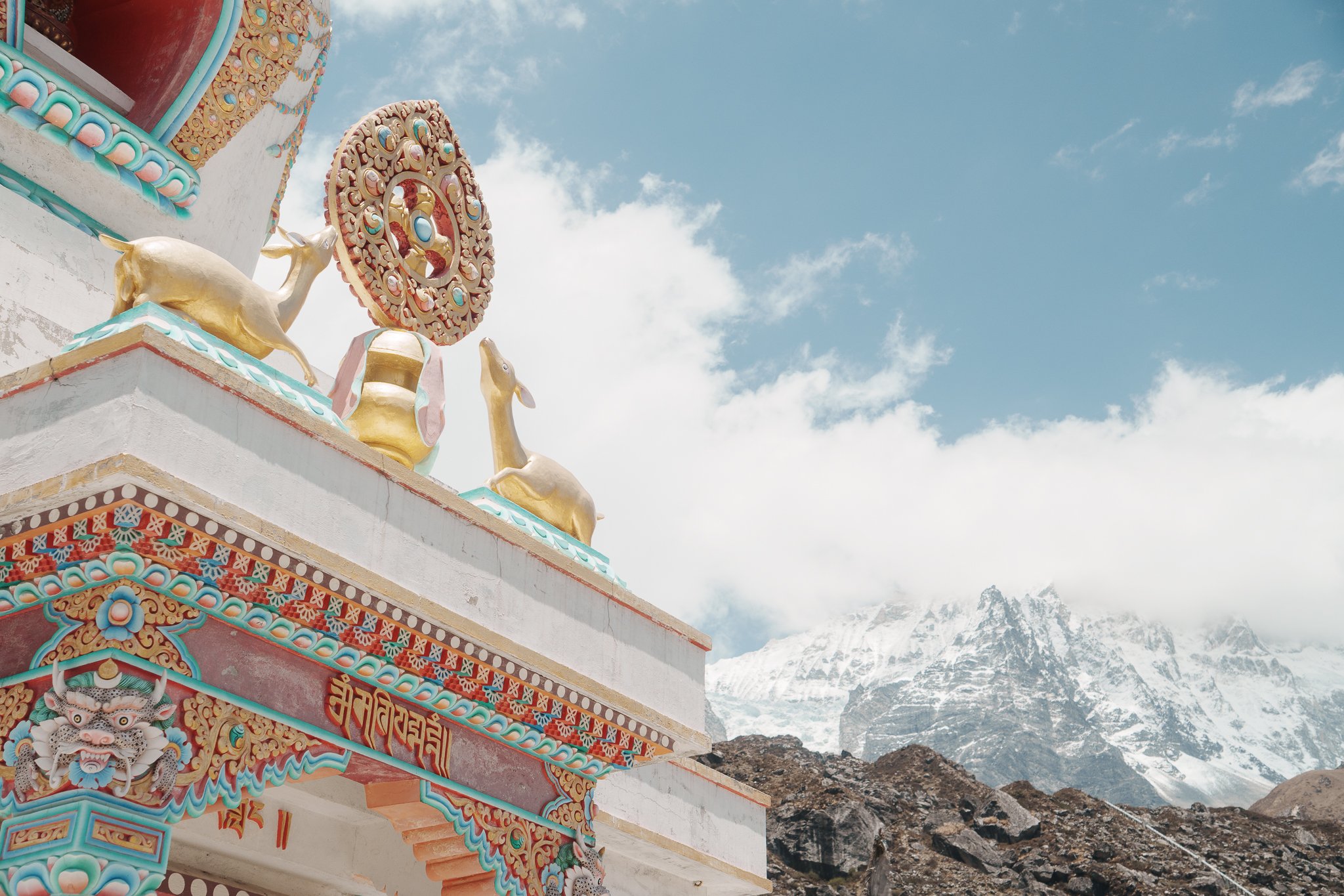 Close-up of a Buddhist temple with colorful details, golden deer statues, and a Dharma wheel, against a backdrop of snow-capped mountains and a partly cloudy sky.