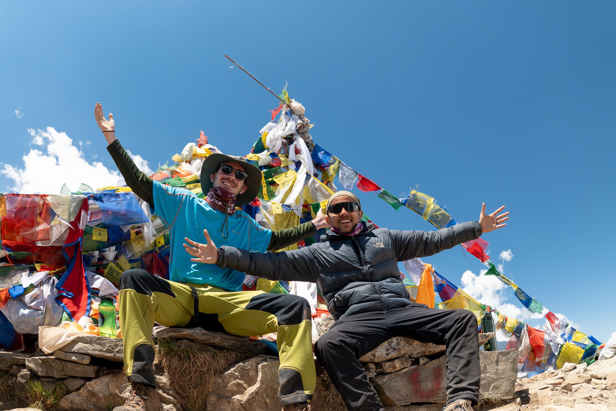 Two men smiling and sitting on rocks at a mountain summit with prayer flags and a bright blue sky behind them.