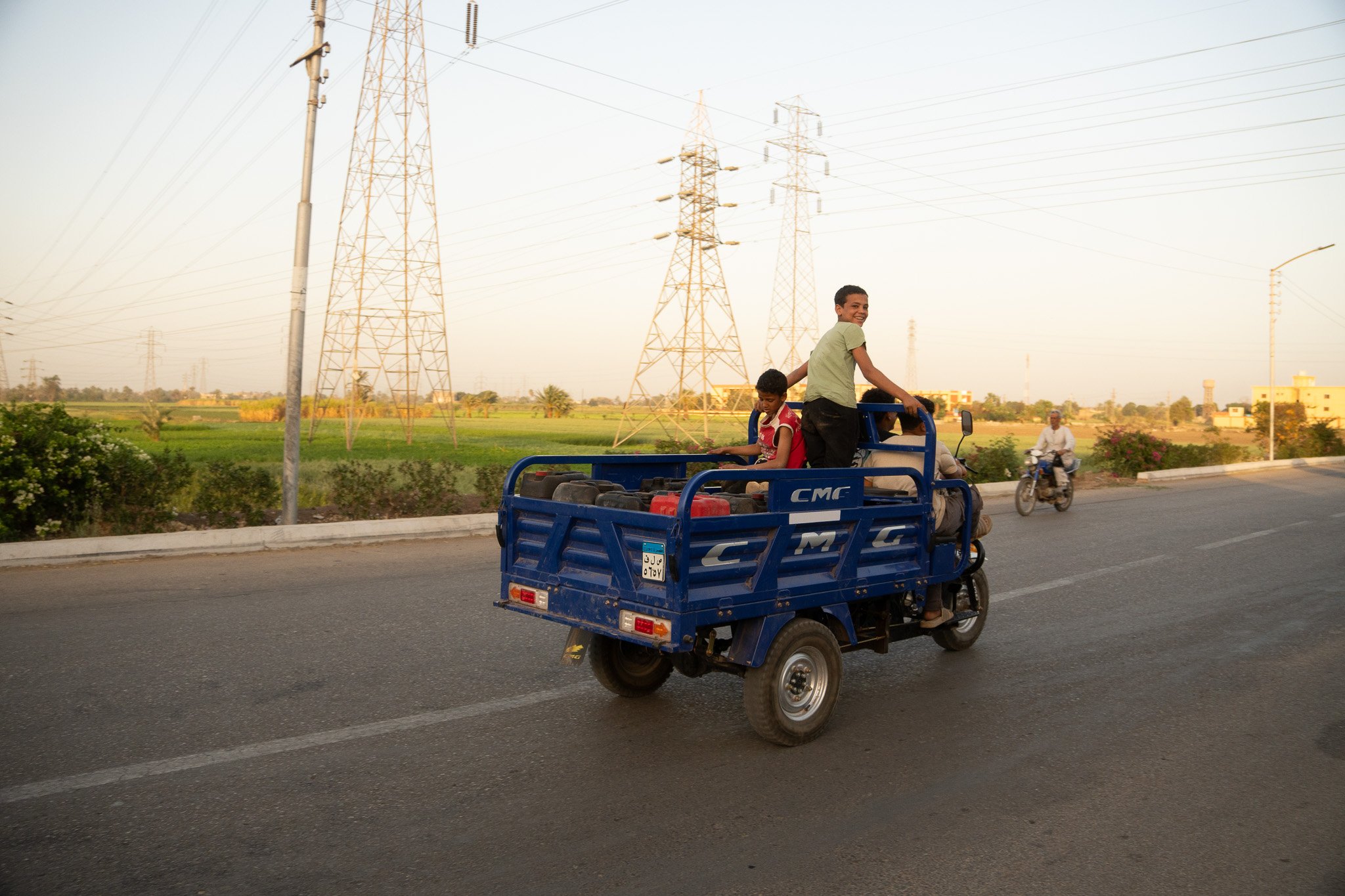 Three children riding in the back of a blue truck on a paved road with power lines and fields in the background.