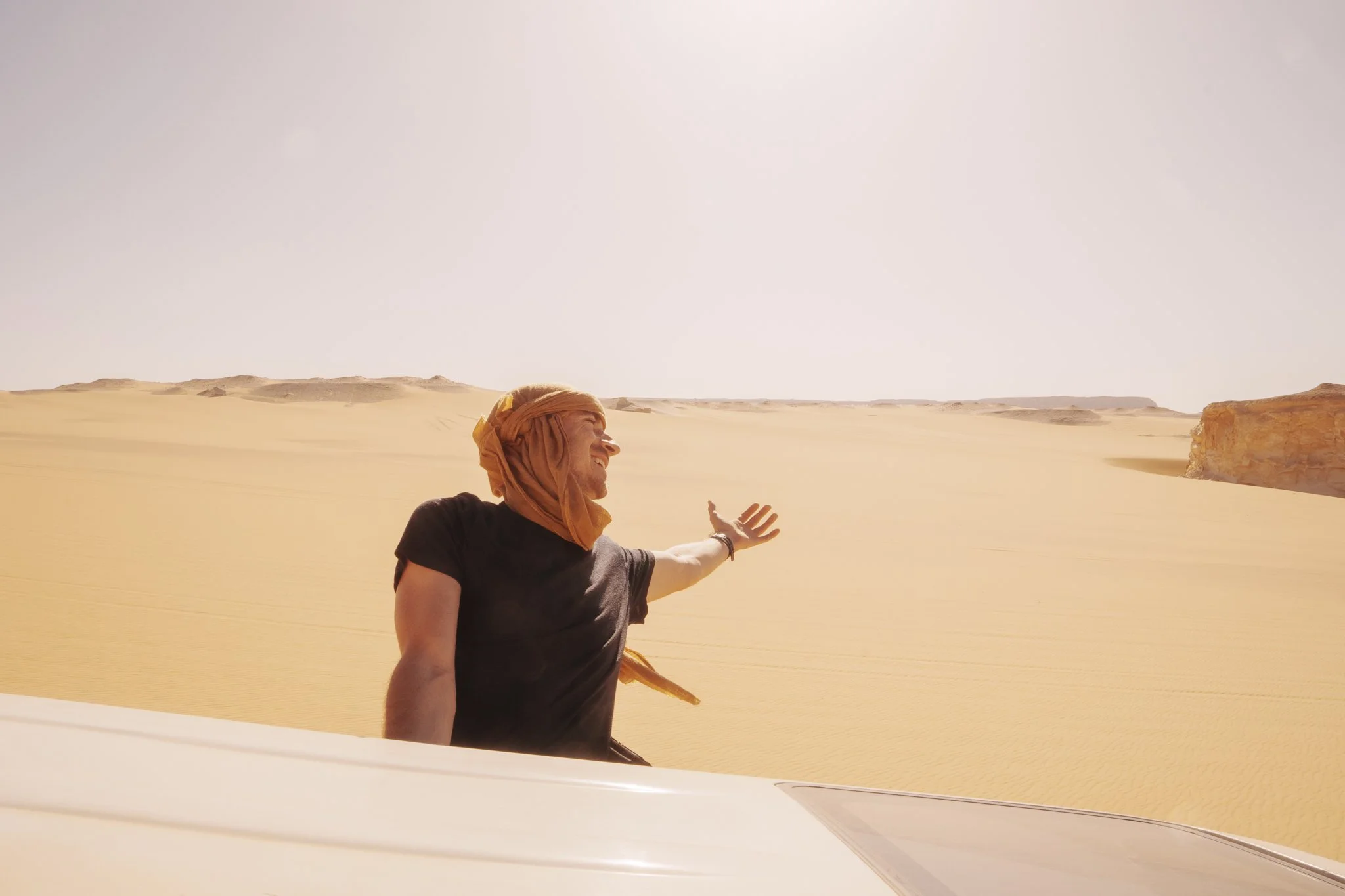 A man wearing a black t-shirt and an orange headscarf standing in a desert with sand dunes and rocky formations in the background, reaching out with his right hand towards the sky and smiling.
