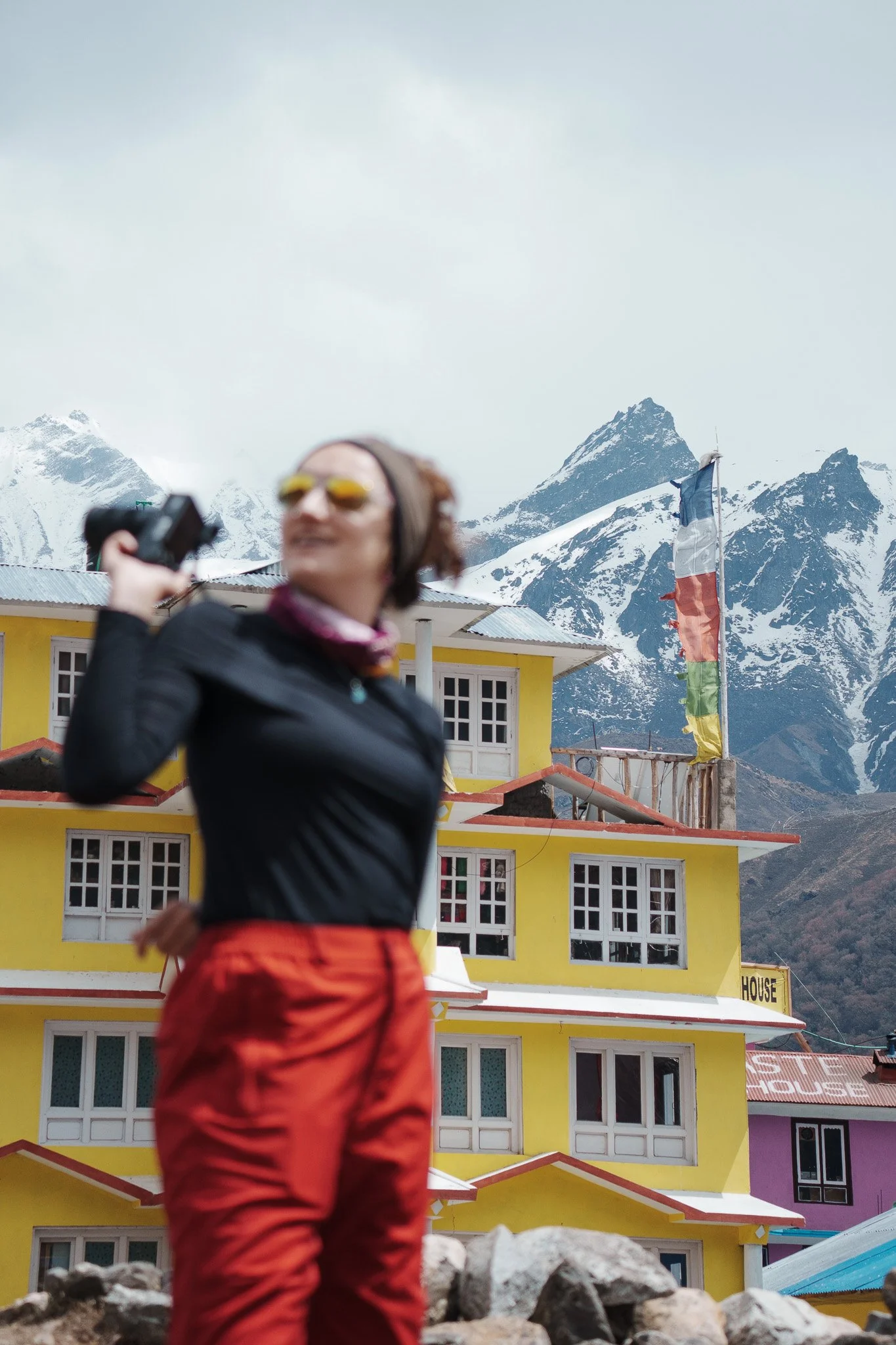 A woman in outdoor clothing holding a camera with snow-capped mountains and a colorful building with a flag in the background.