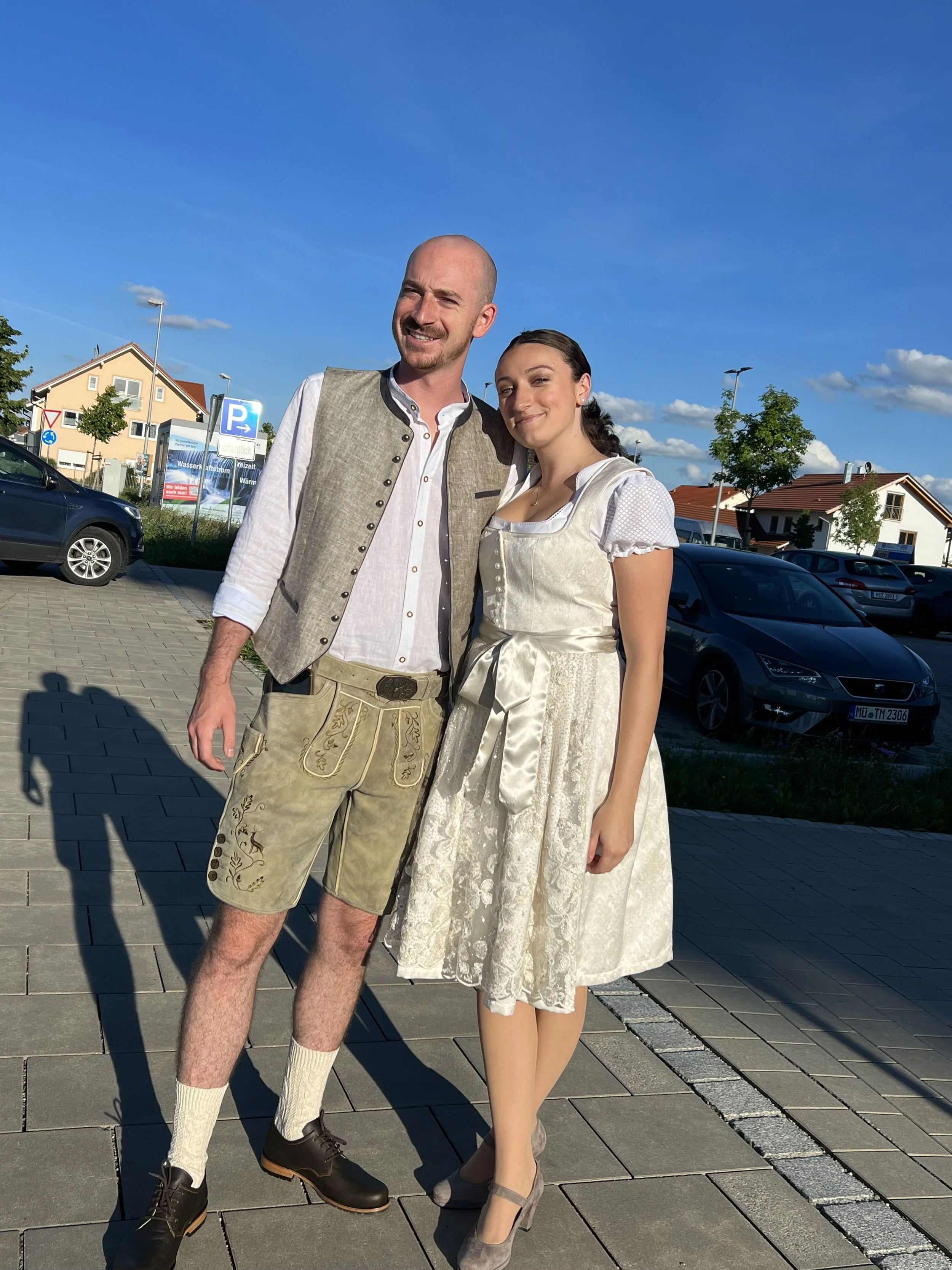A man and woman dressed in traditional Bavarian clothing standing outdoors on a sunny day, smiling at the camera. The man wears khaki Lederhosen, a white shirt, and a vest, while the woman wears a cream-colored dirndl with lace details and heels.