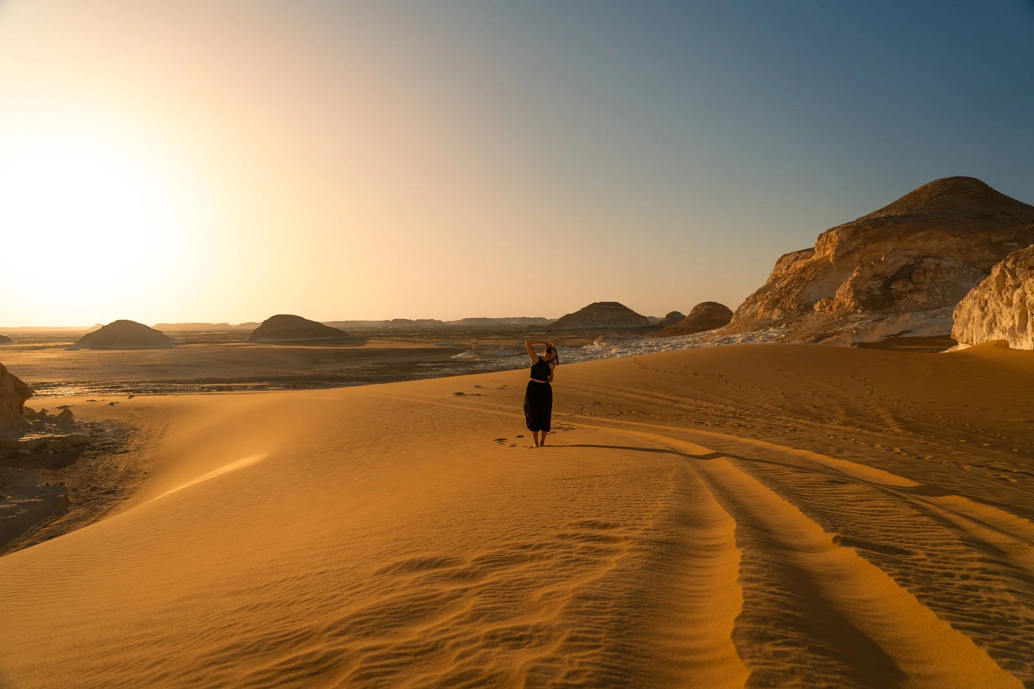 A person standing on sandy dunes at sunset, with large rocky formations and hills in the background.