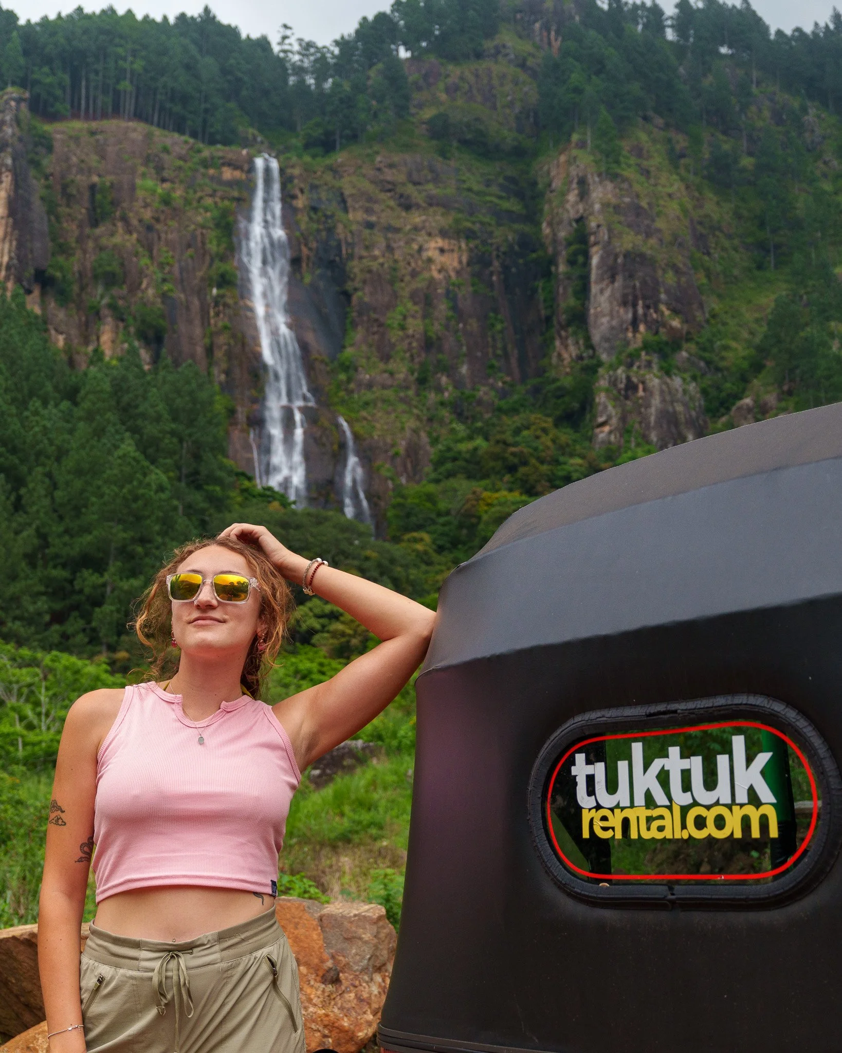 A woman with sunglasses and a pink tank top standing outdoors near a large black tent with a logo reading tuktuk rental.com, in front of a lush green forest and a waterfall on a mountainous landscape.