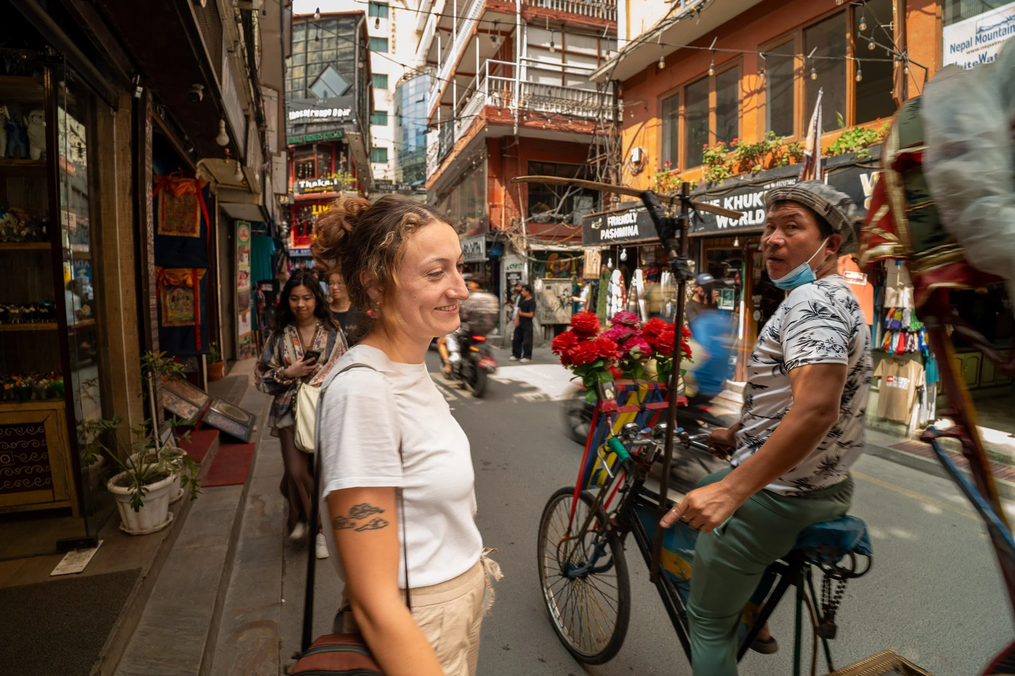 Two women standing and talking on a busy street in Nepal with shops and people in the background. One woman is smiling, and the other is riding a bicycle with flowers attached to it.
