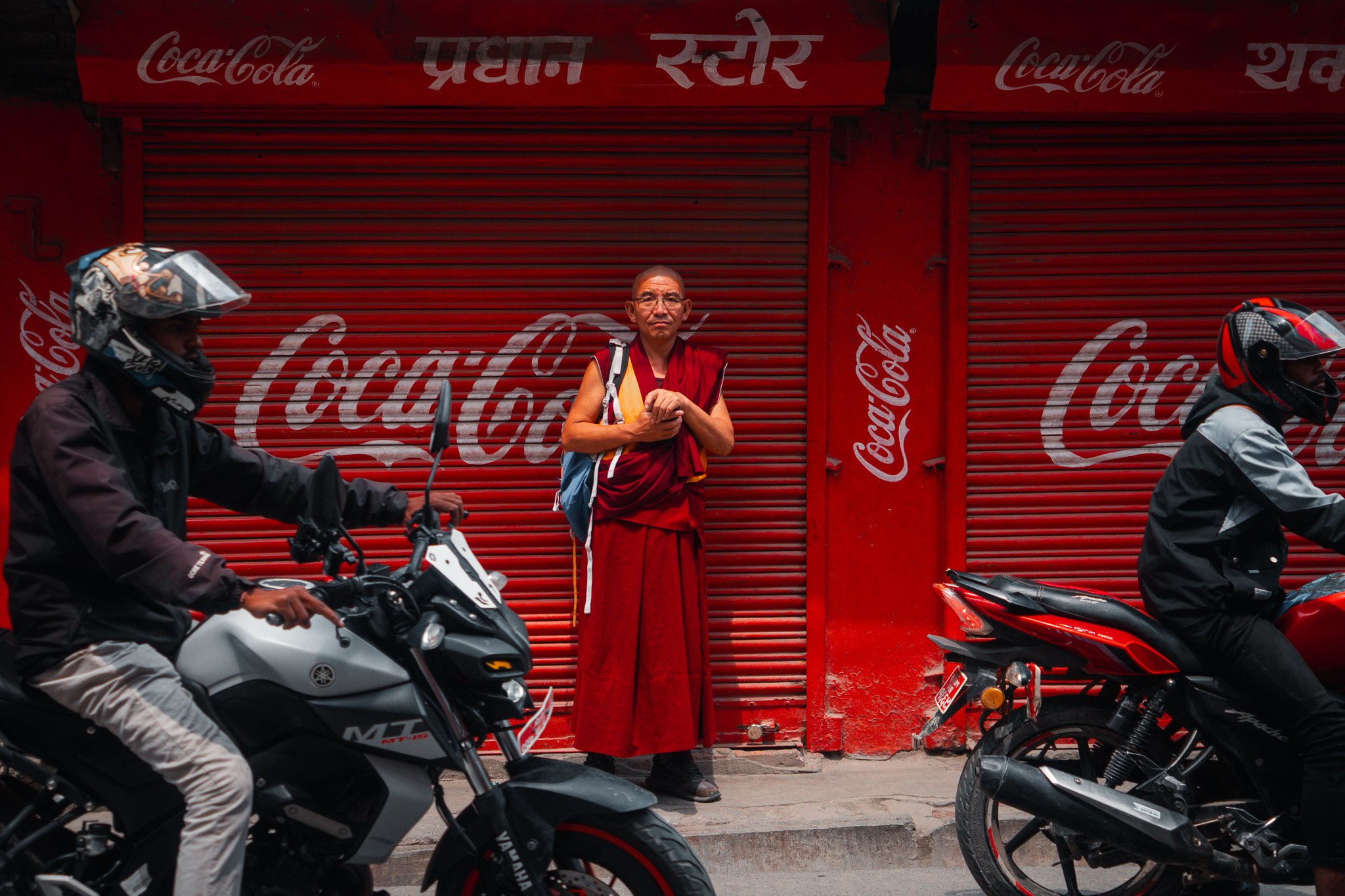 A monk in red robes standing with hands clasped in front of a closed red shop with Coca-Cola branding, while two motorcyclists pass by in opposite directions on a busy street.