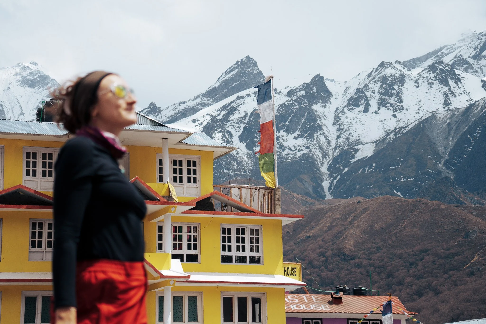 Blurred woman in a black jacket and red pants walking past a yellow house with a mountain range in the background.