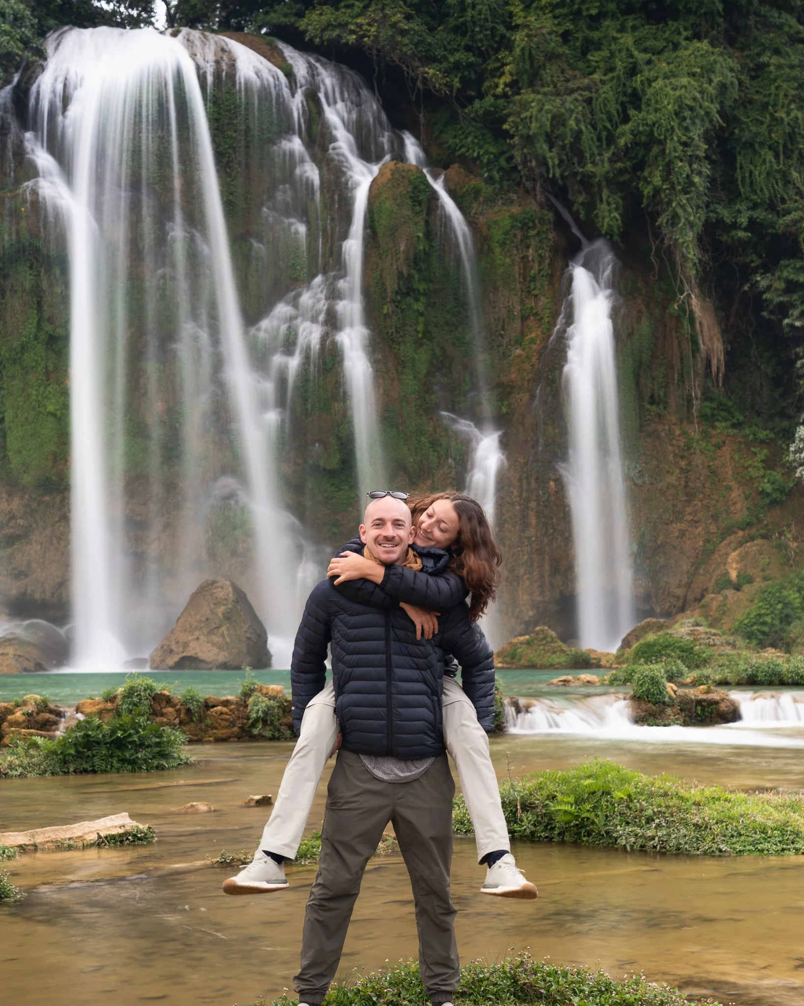 A man carrying a woman on his back in front of a large waterfall surrounded by green trees.