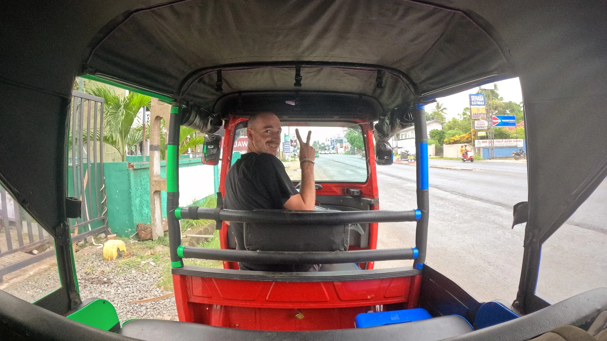 A man sitting in the back of a red tuk-tuk, smiling and making a peace sign, on a city street during daytime.