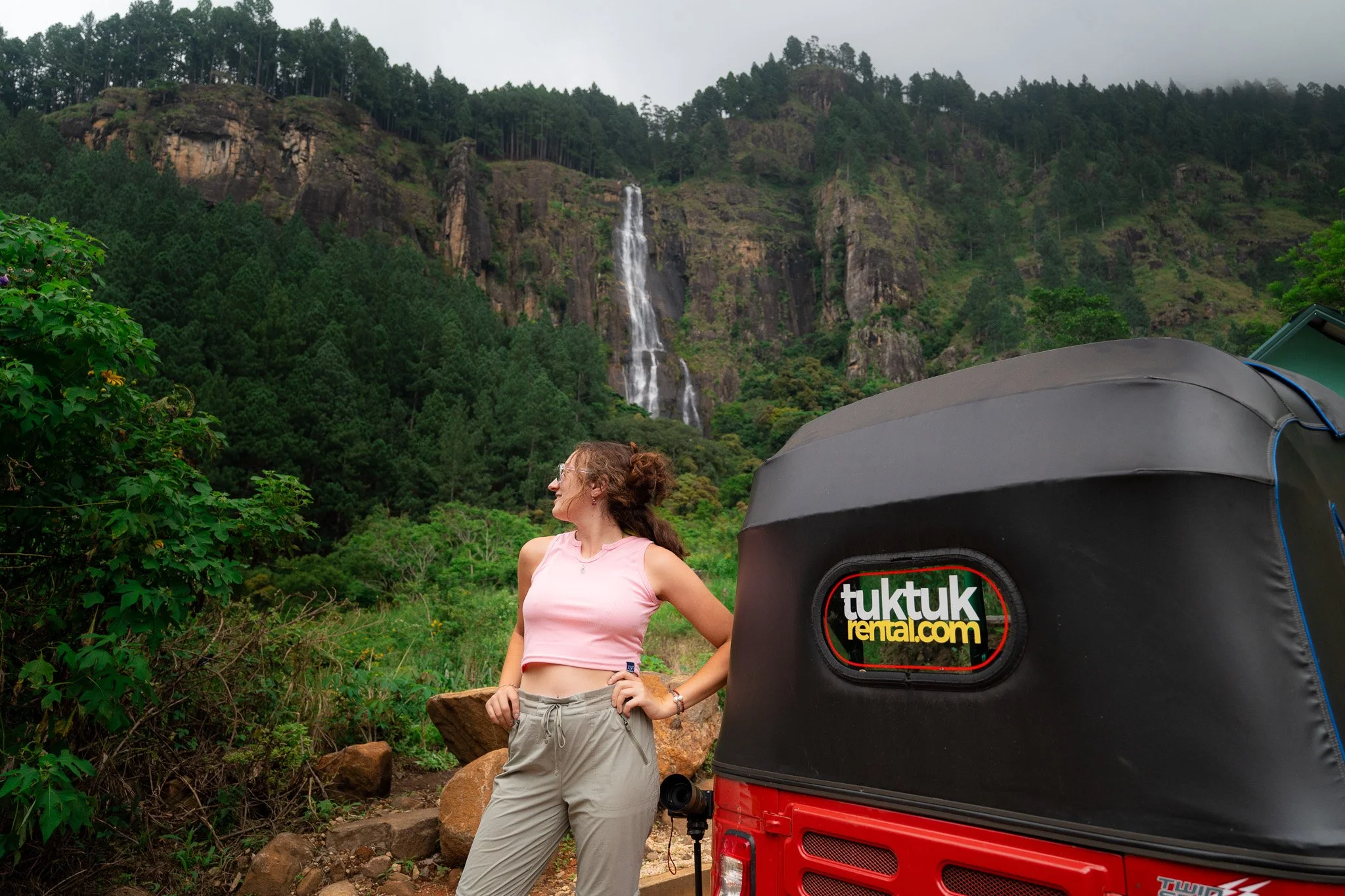 A woman with curly hair wearing sunglasses, a pink sleeveless top, and beige pants, standing outdoors in front of a lush green landscape with a waterfall cascading down a mountain in the background. A tuk tuk with a black cover and a logo for tuk tuk