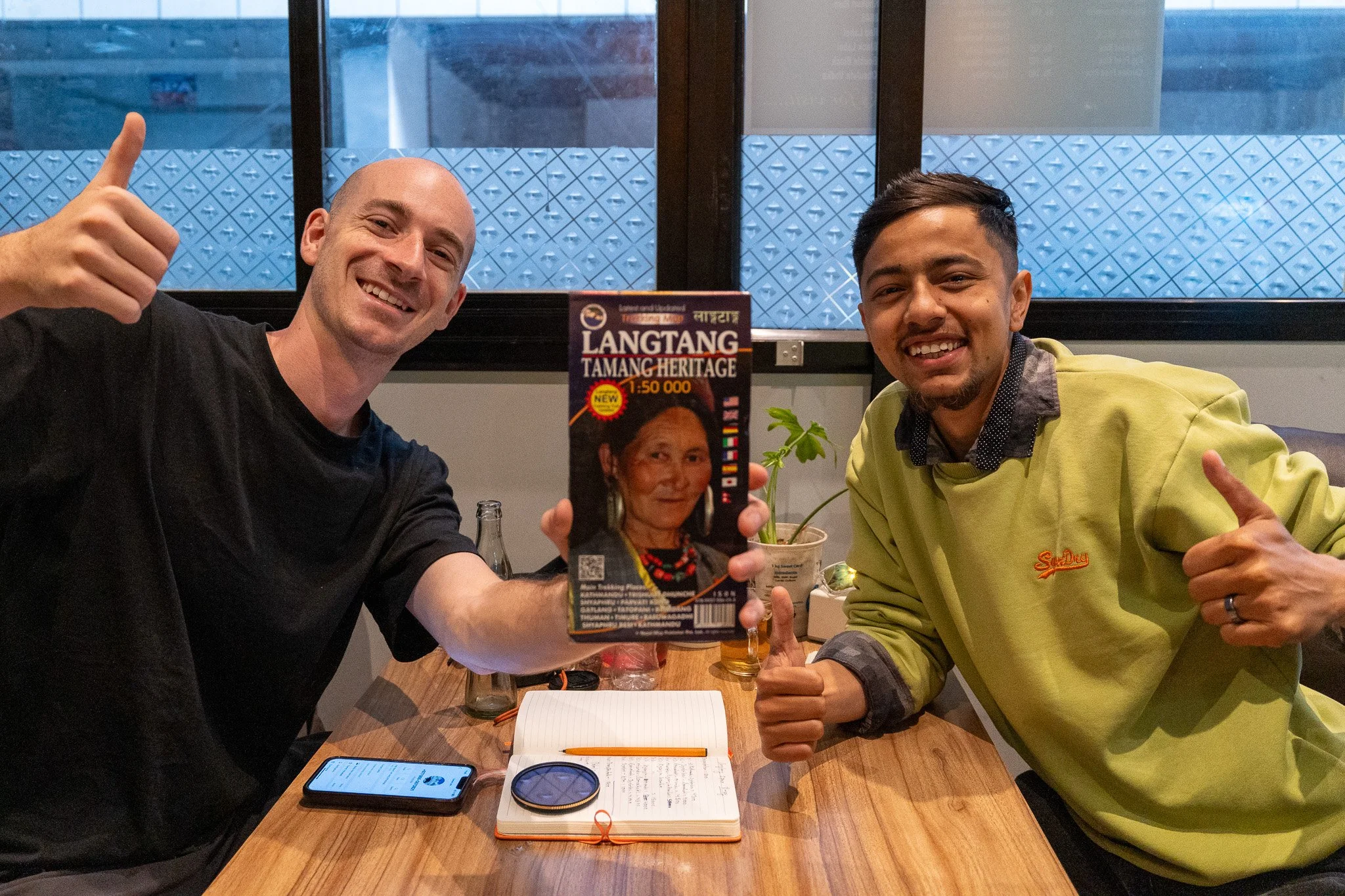 Two men smiling at the camera, giving thumbs up, sitting at a table with a pack of Langtang Tamang Heritage model, notebooks, and drinks, inside a restaurant.