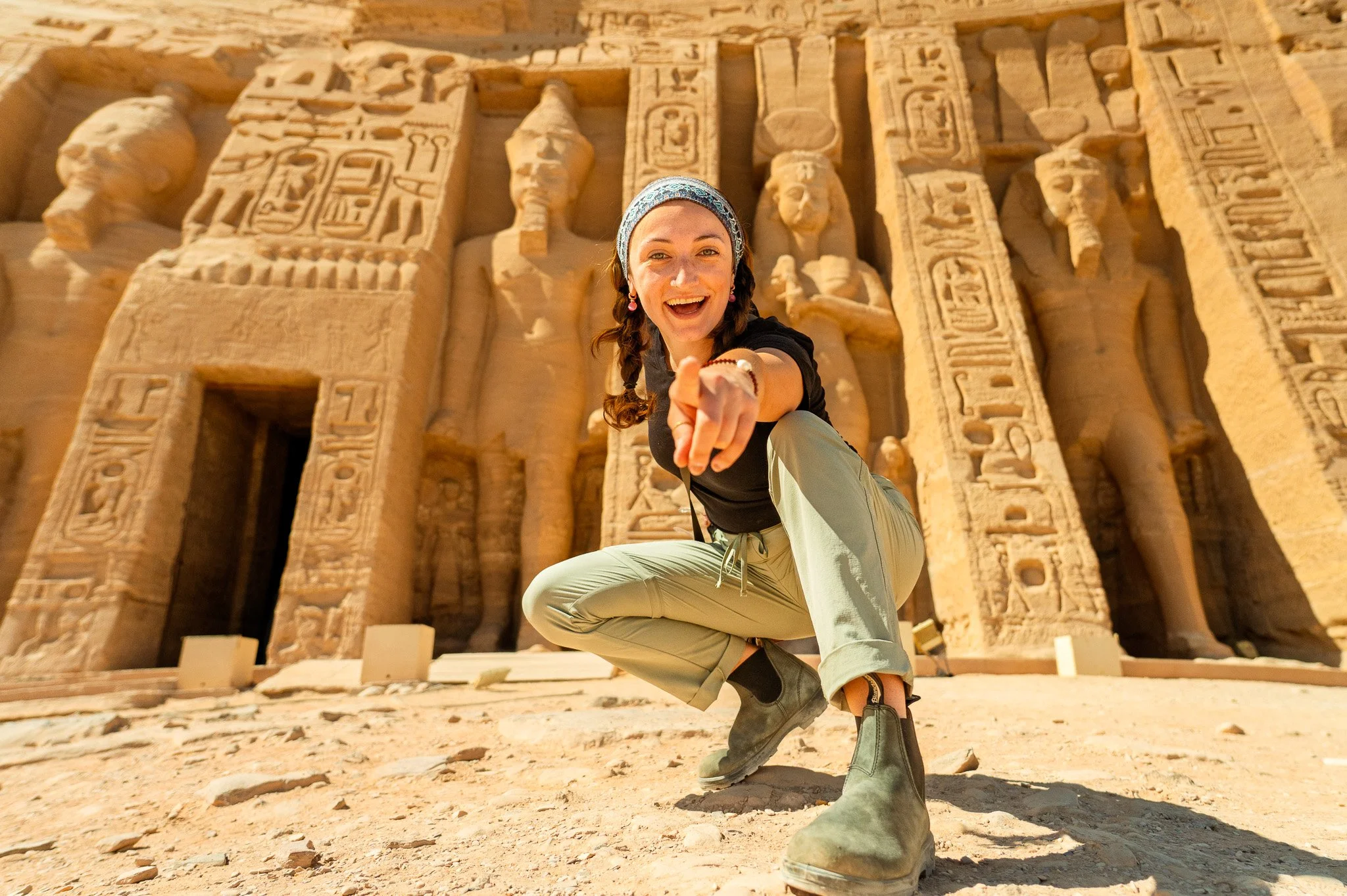 A woman crouching and smiling, pointing towards the camera, in front of an ancient Egyptian temple with hieroglyphics and statues carved into the stone walls.