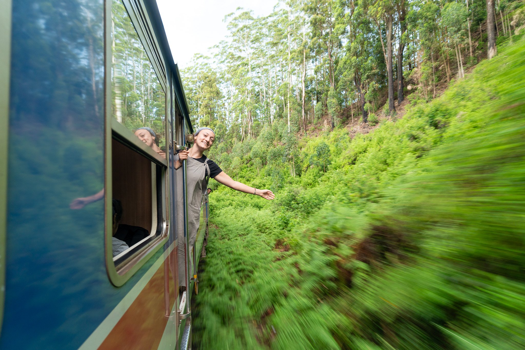 A woman smiling and leaning out of a train window, waving her hand as the train travels through a lush green forest.