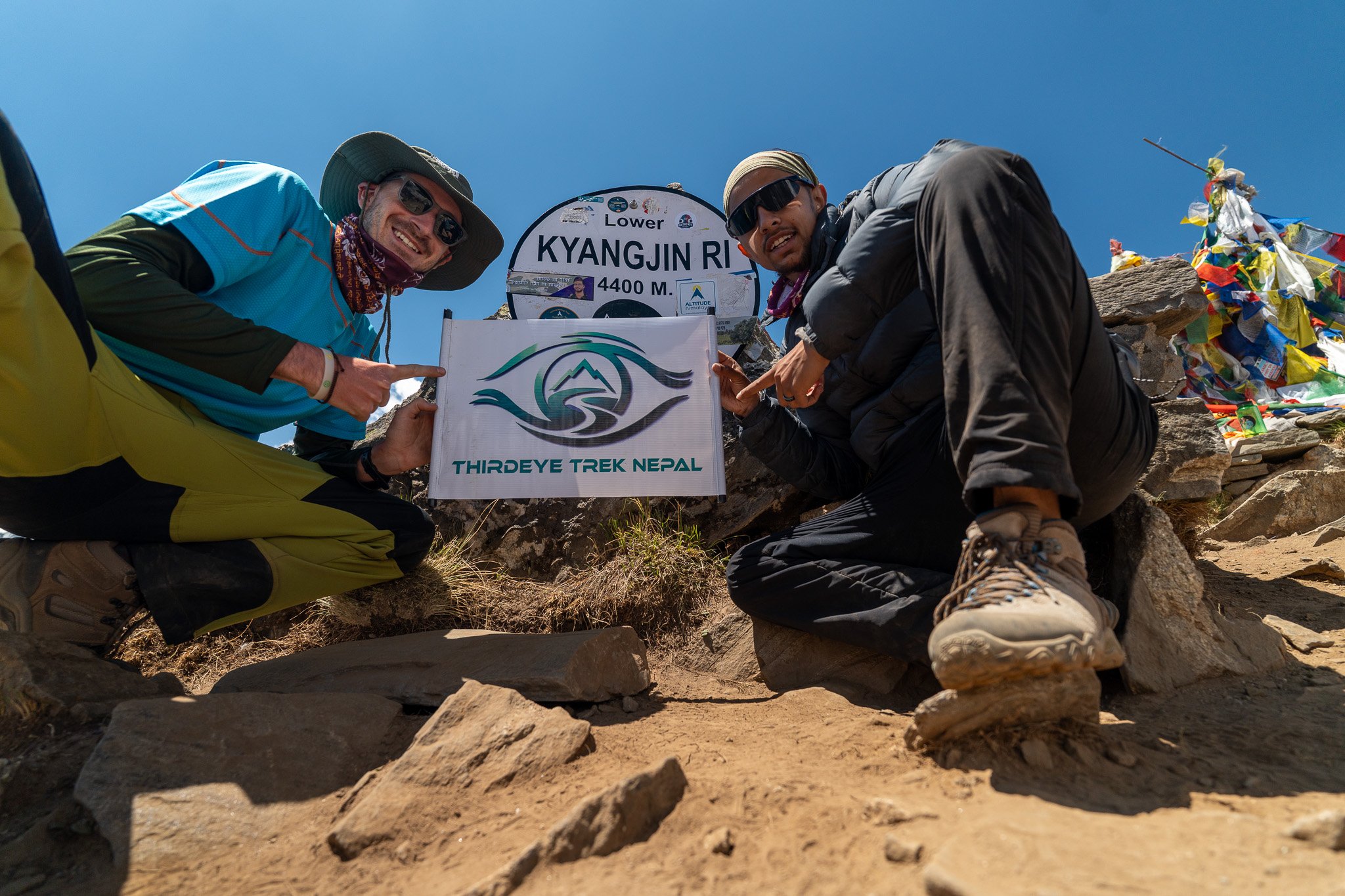 Two male hikers at the summit of Kyangjin Ri in Nepal, holding a sign that reads "Thirdeye Trek Nepal" with a mountain logo, surrounded by rocks and prayer flags, under a clear blue sky.