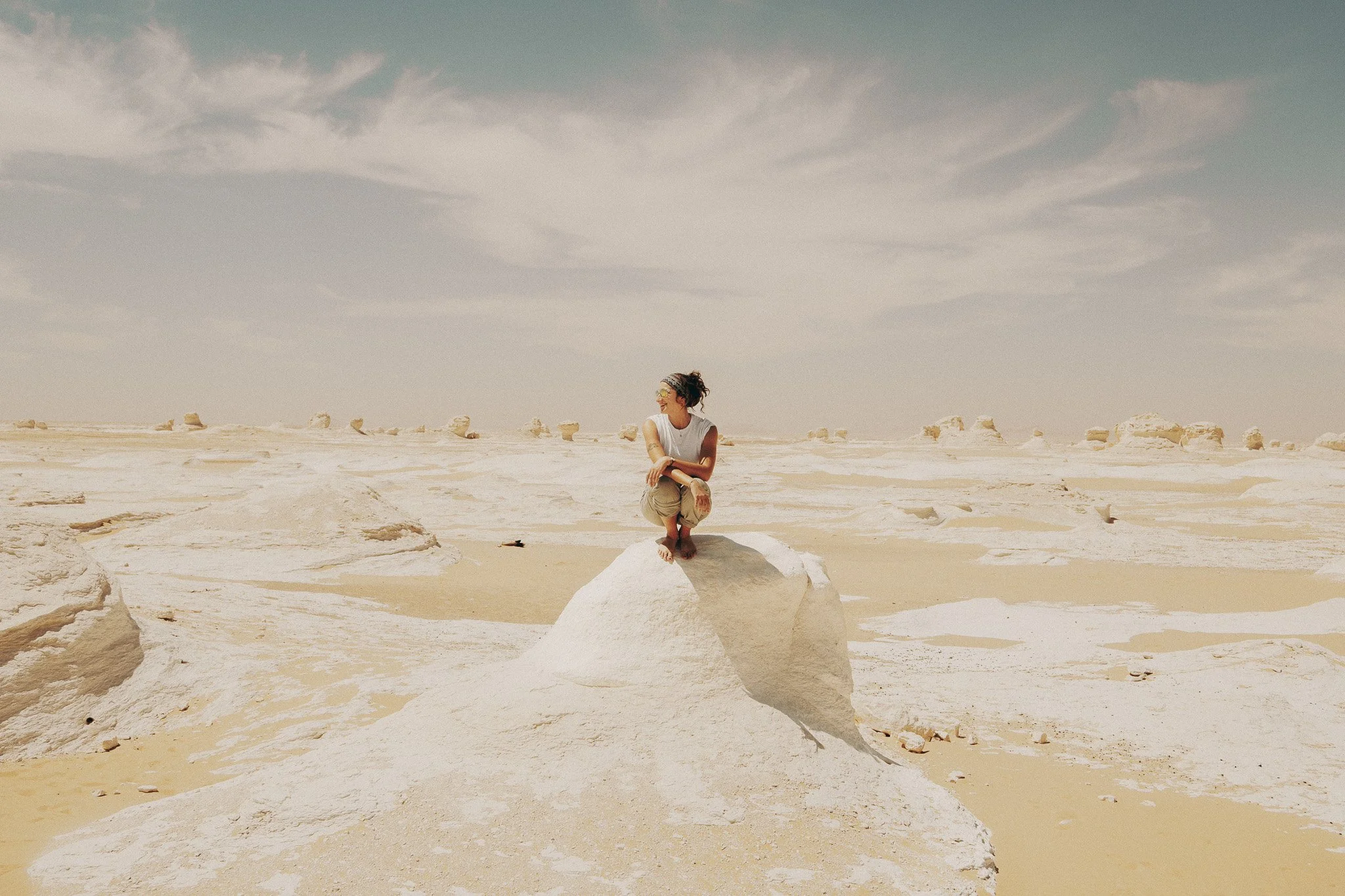 A woman squatting on a large white rock in a desert with beige sands and scattered white rocks, under a partly cloudy sky.