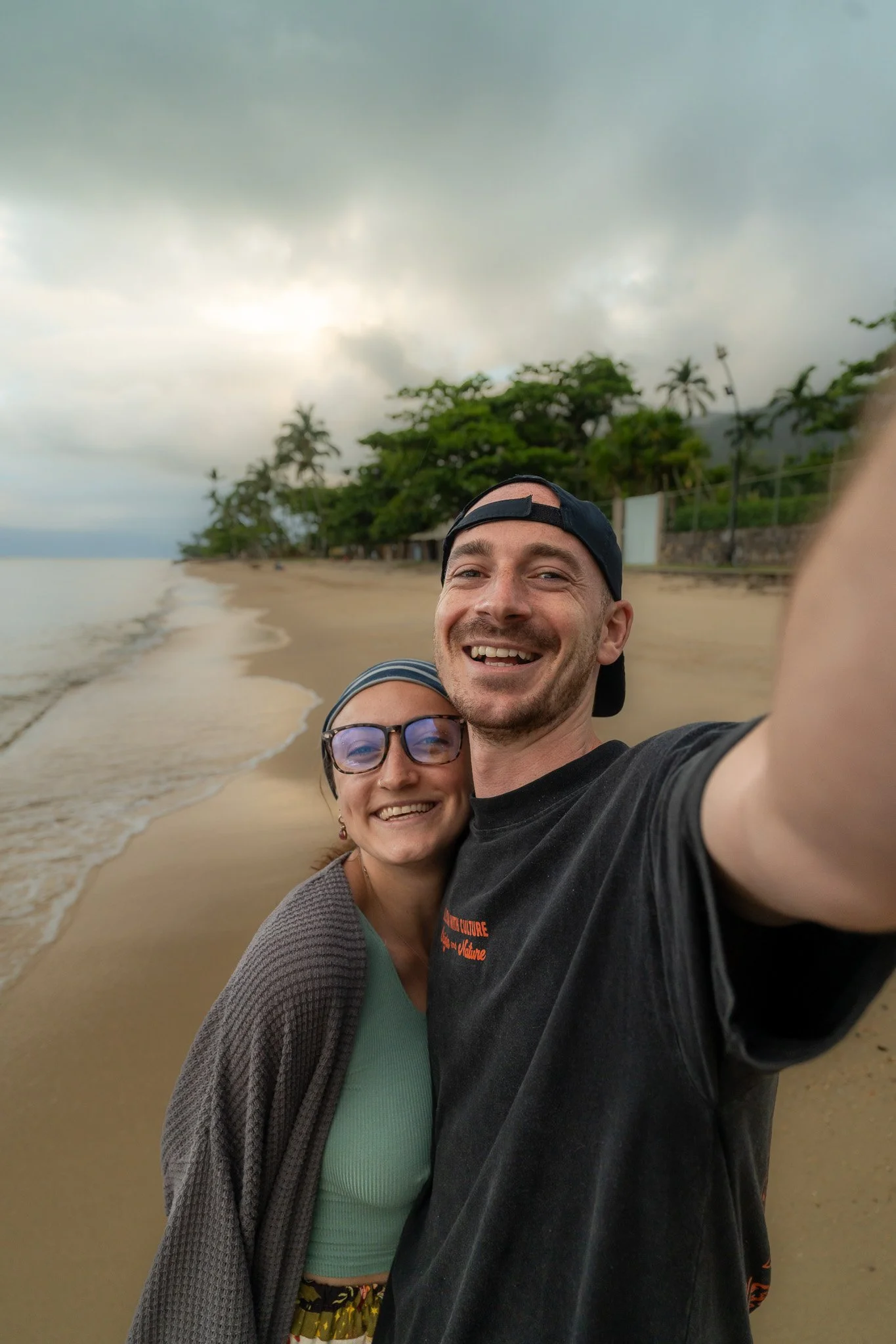 A man and woman smiling for a selfie on a beach with sand, ocean, trees, and cloudy sky in the background.